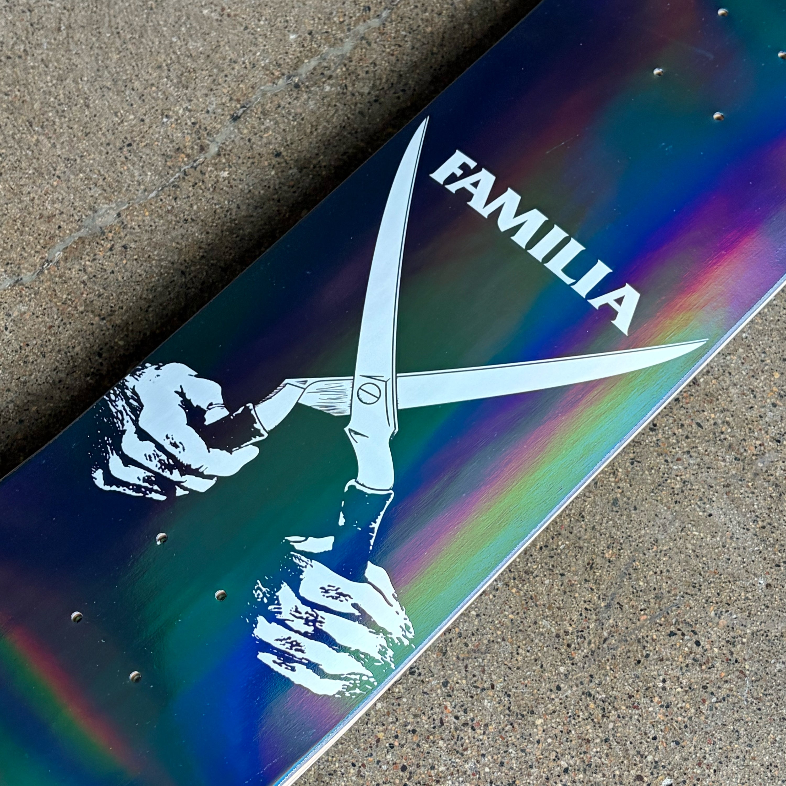 A close-up of a skateboard deck featuring a holographic iridescent finish with a white graphic of a hand holding scissors and the word 'FAMILIA' printed in white.