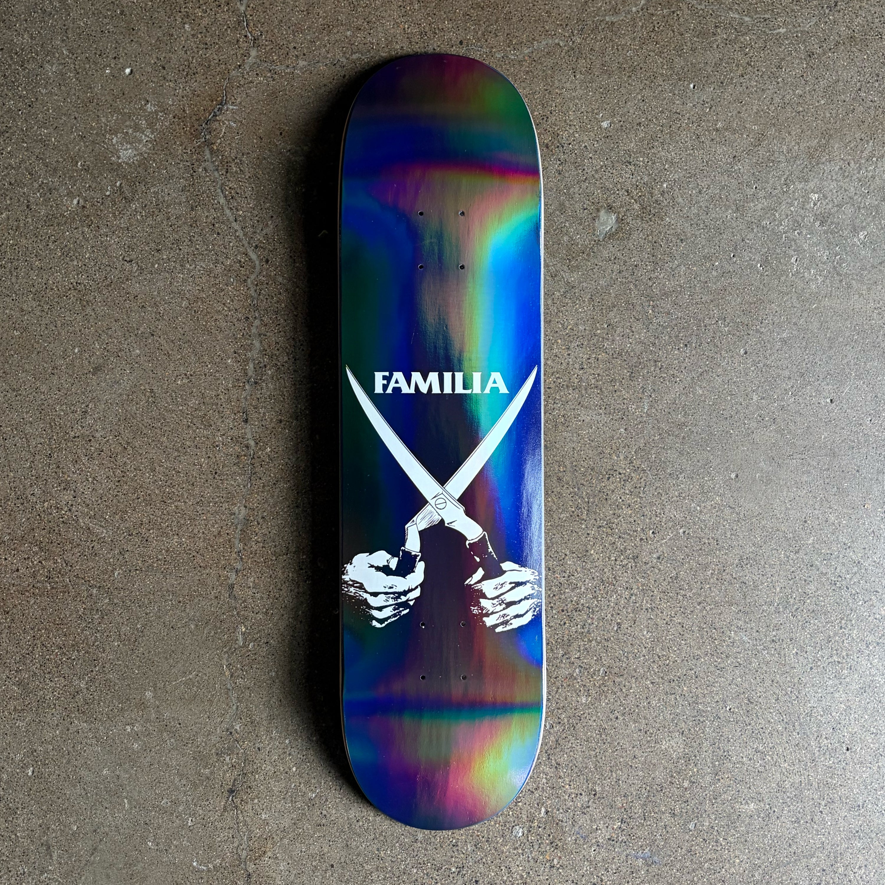 Full image of a skateboard deck with art of hands holding a pair of shears and the  'Familia' in white. Features an iridescent shine. Photographed from above on a concrete floor.