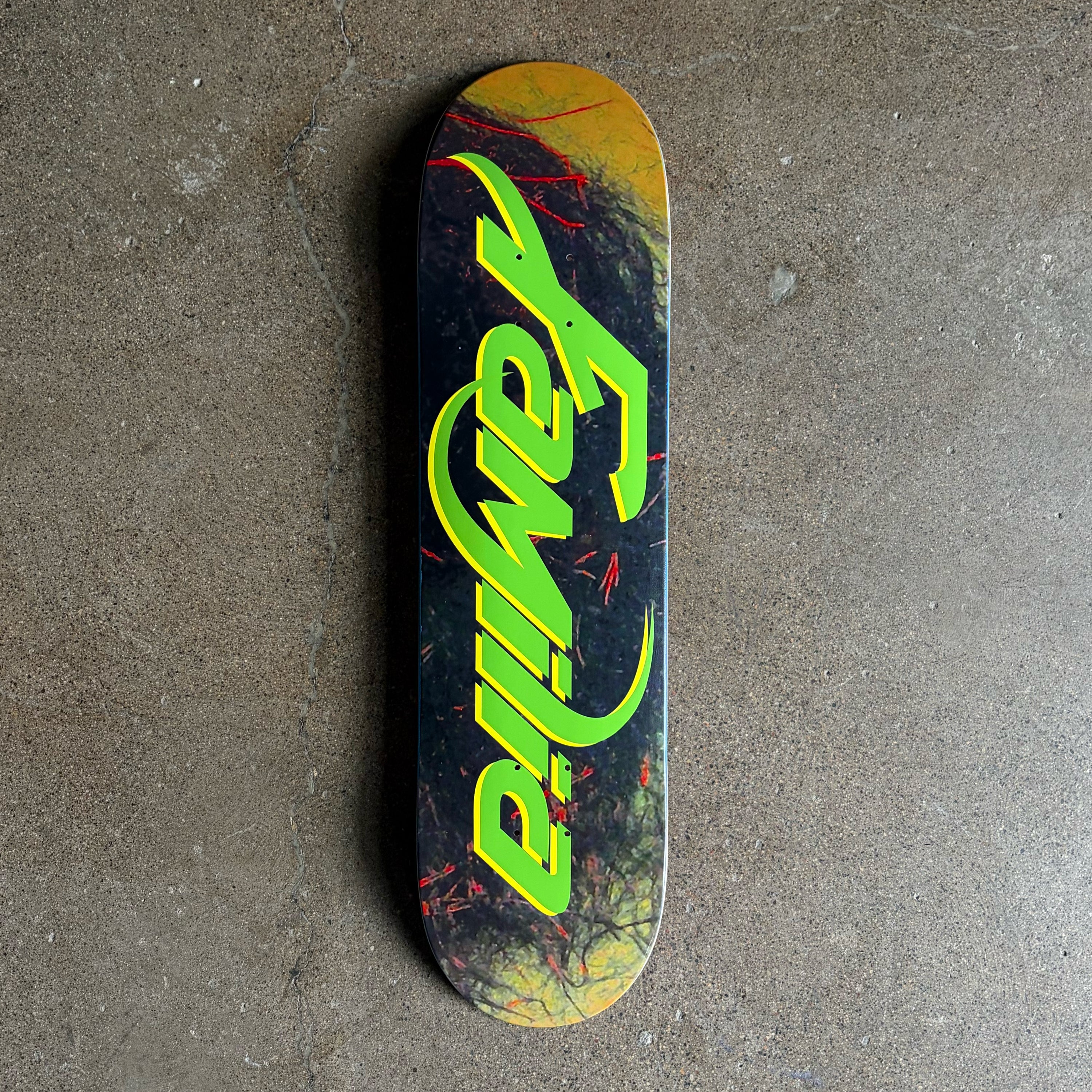 A skateboard deck with a dark graphic background, yellow-orange distressed edges, and a large neon green 'Familia' logo, photographed from above on a concrete floor.