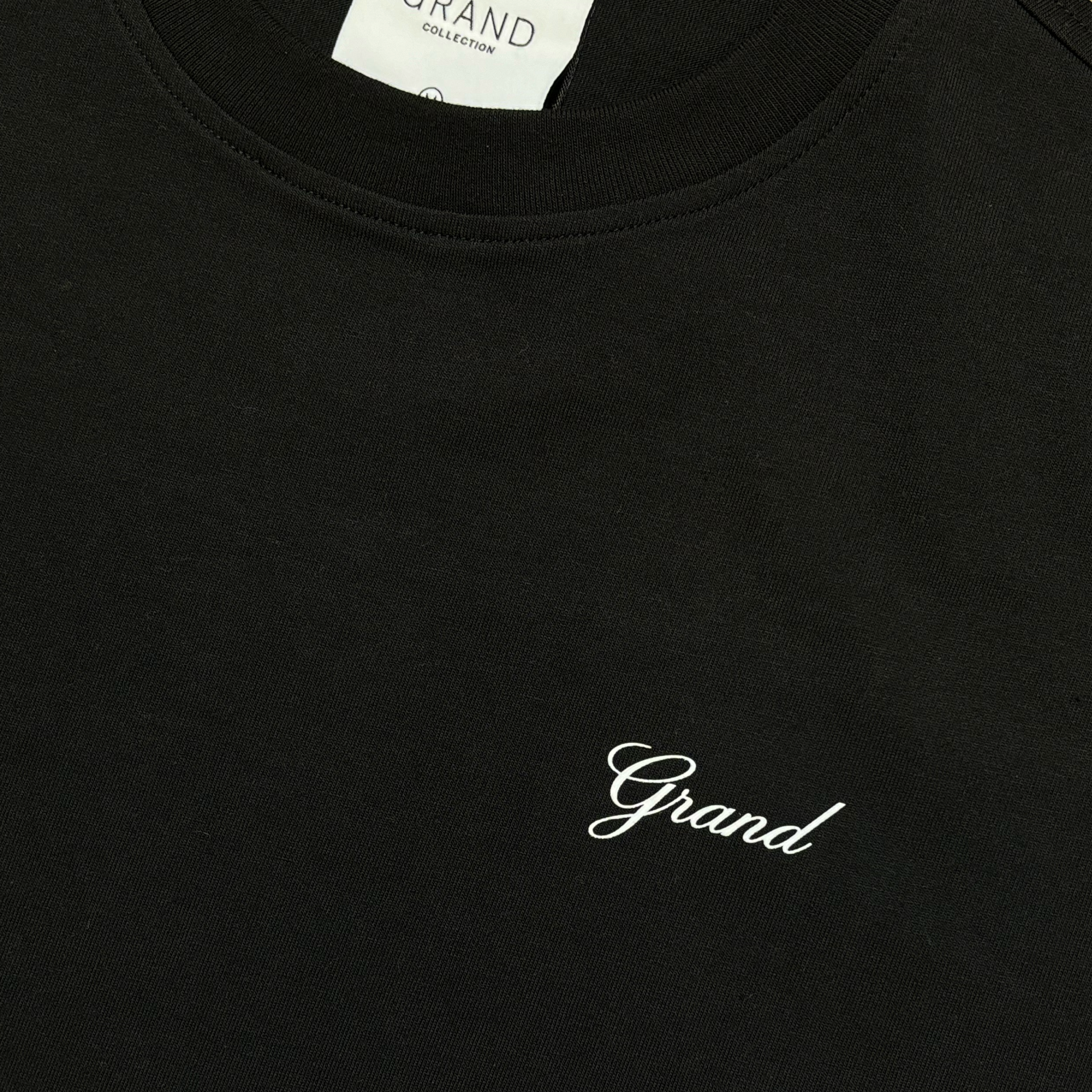 CLOSE UP ANGLE OF BLACK T-SHIRT, GRAPHIC IN CURSIVE FONT THAT READS GRAND ON LEFT CHEST