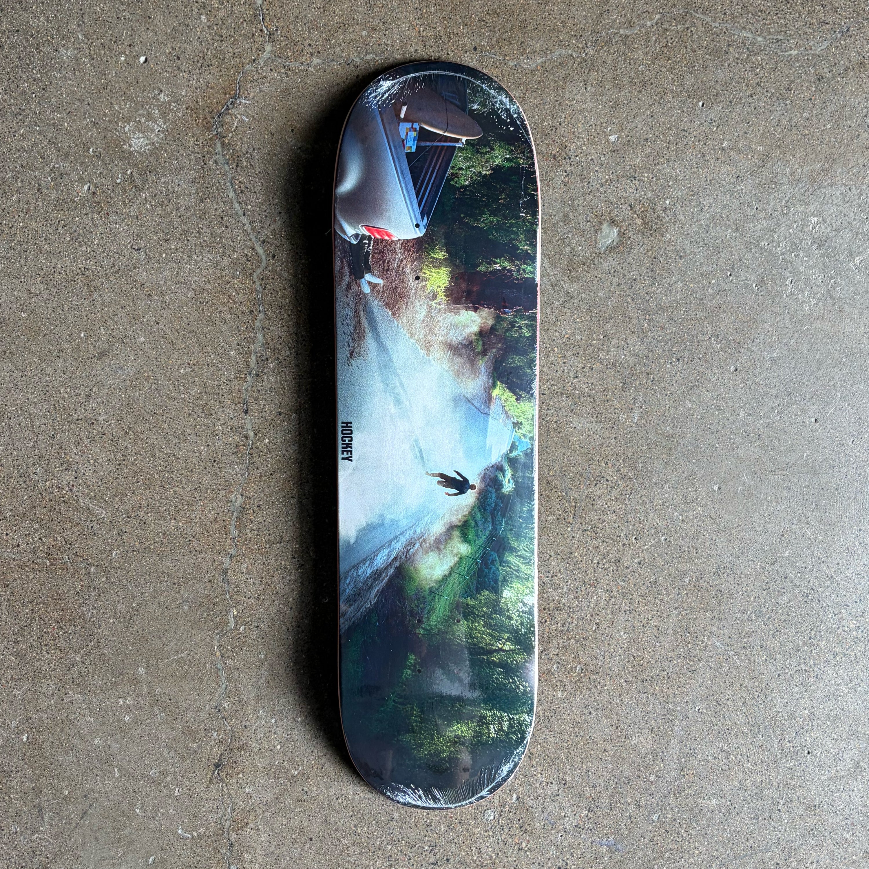 A skateboard deck with a scenic waterfall and forest graphic, photographed from directly above on a textured concrete floor.