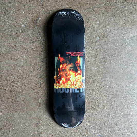 A Hockey skateboard deck wrapped in protective plastic, featuring a black background with a central graphic of a face engulfed in flames, placed on a concrete floor.