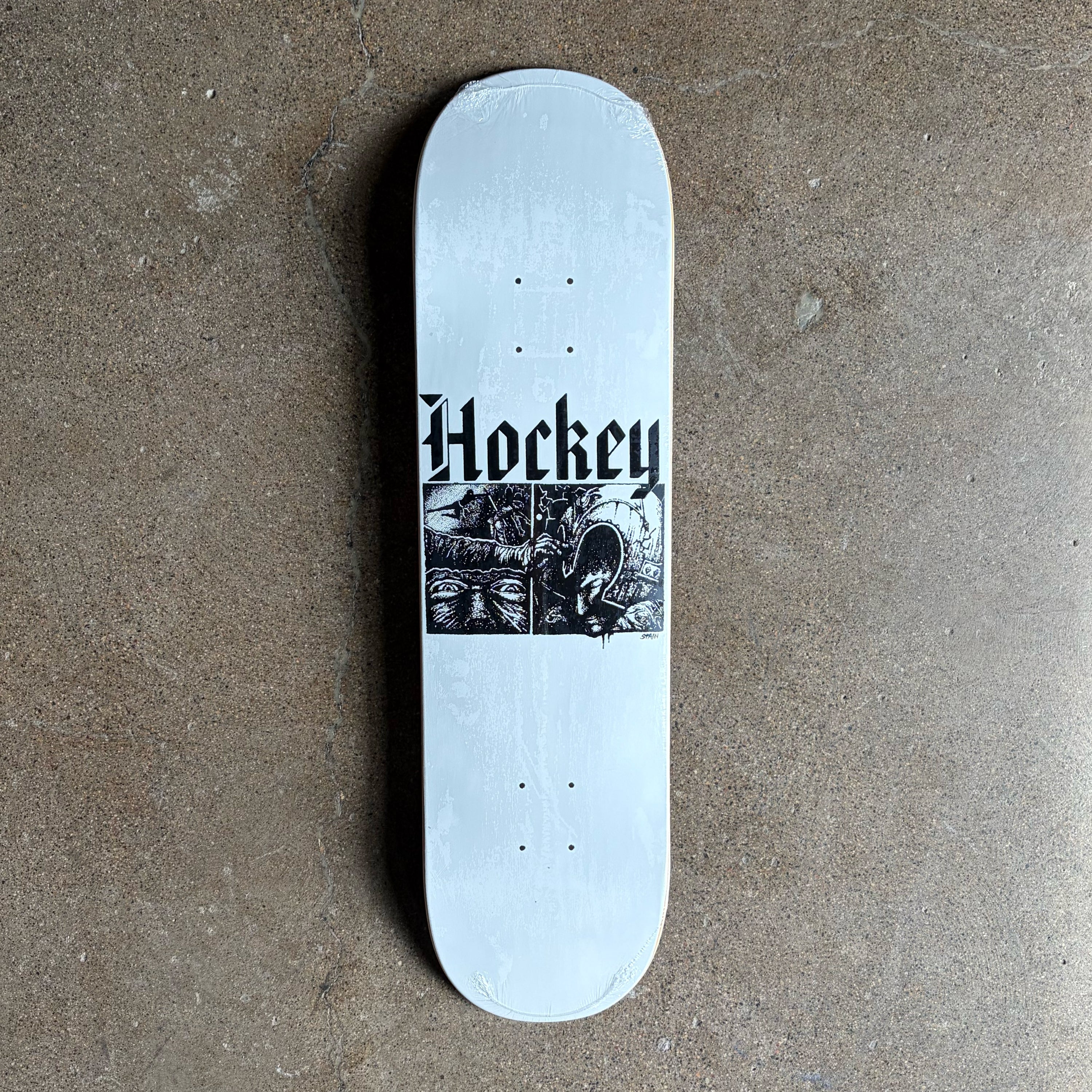 A white skateboard deck with a black graphic print featuring the word 'Hockey' in a gothic font and a black-and-white illustration, photographed from above on a textured concrete floor.