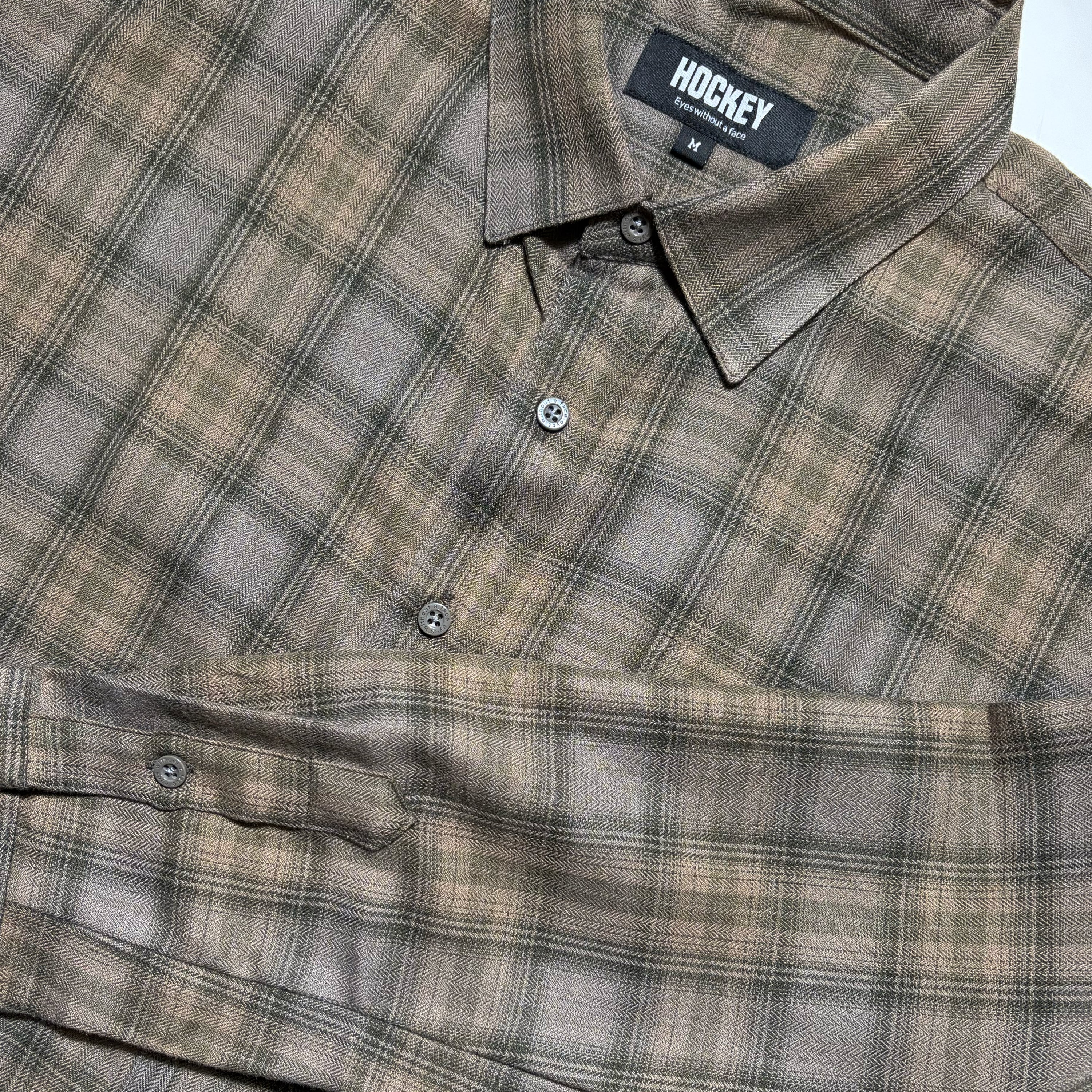 A close-up of a plaid button-up shirt by HOCKEY, featuring a muted brown, beige, and gray pattern. The brand label on the collar reads 'HOCKEY'.