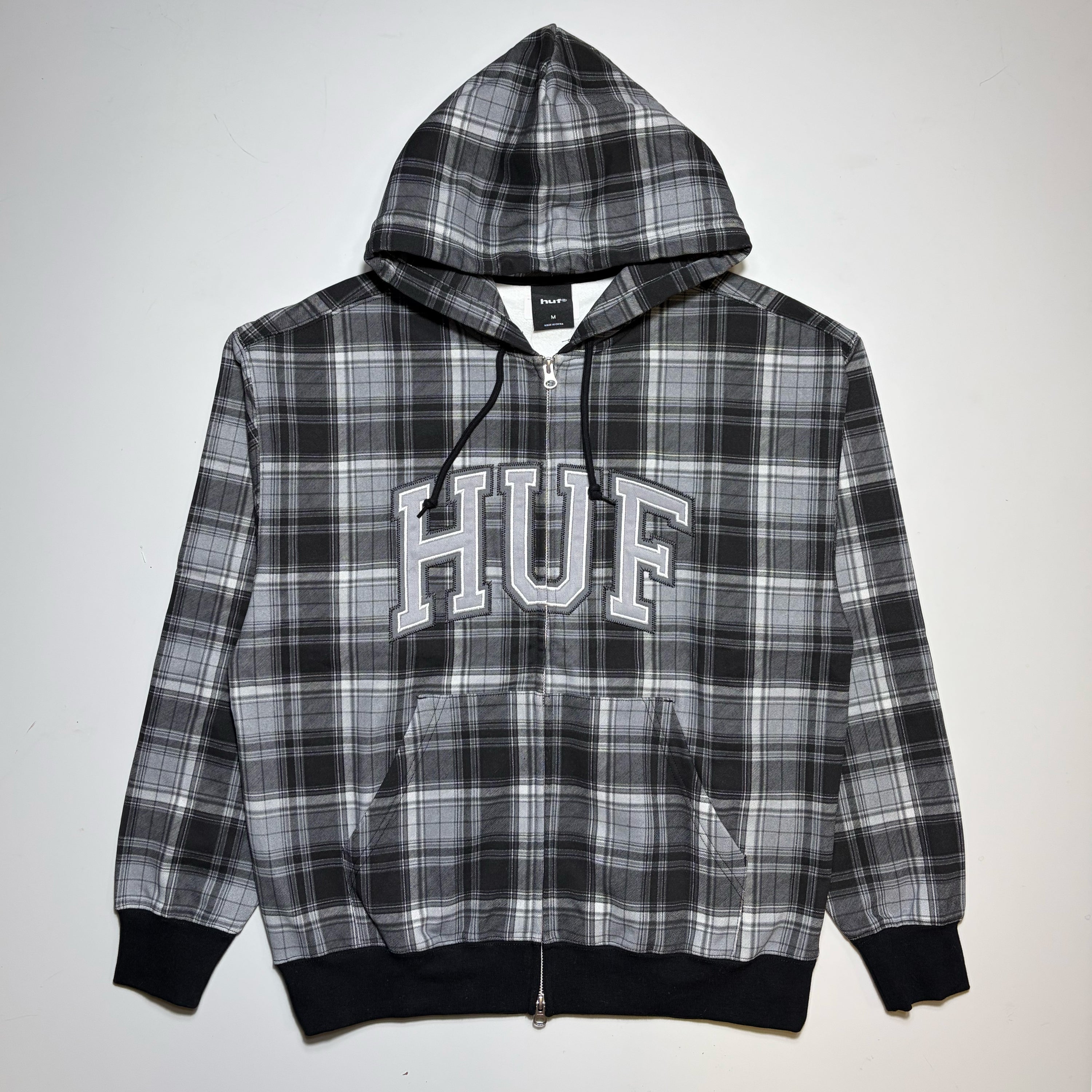 A black, white, and gray plaid zip-up hoodie with 'HUF' printed in white varsity letters across the chest, displayed flat against a light gray background.