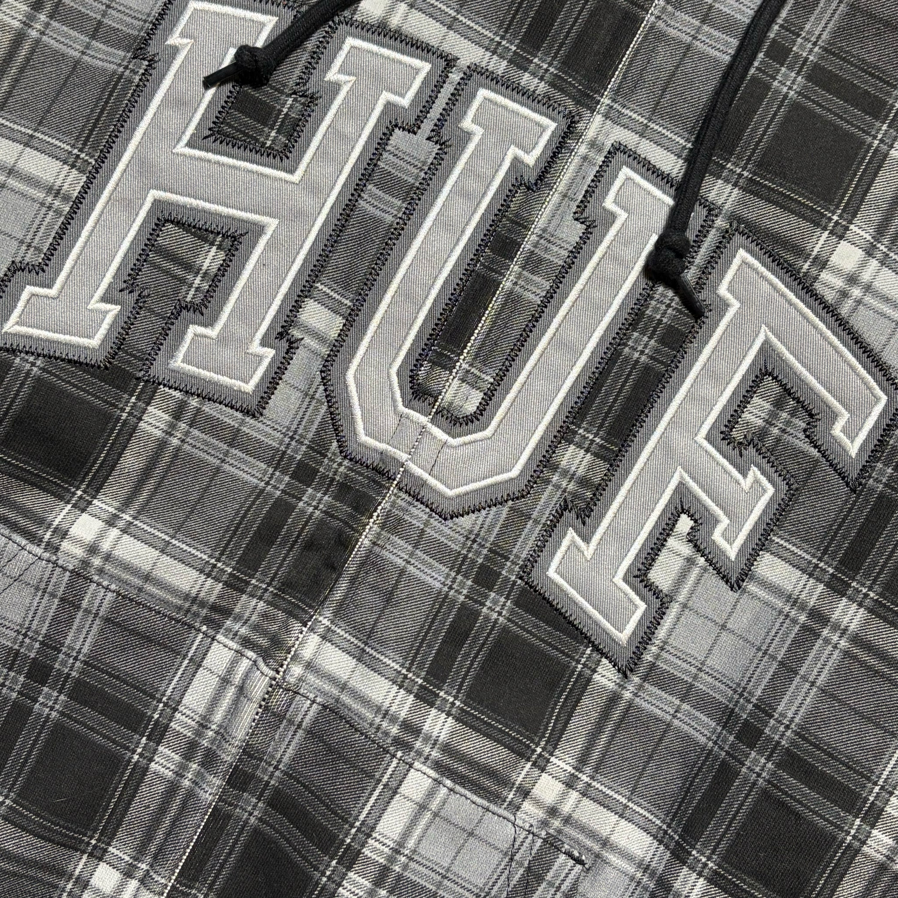 Close-up of a gray and black plaid shirt featuring large embroidered varsity-style letters 'HUF' outlined in white, with black drawstrings visible at the top.