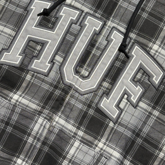 Close-up of a gray and black plaid shirt featuring large embroidered varsity-style letters 'HUF' outlined in white, with black drawstrings visible at the top.