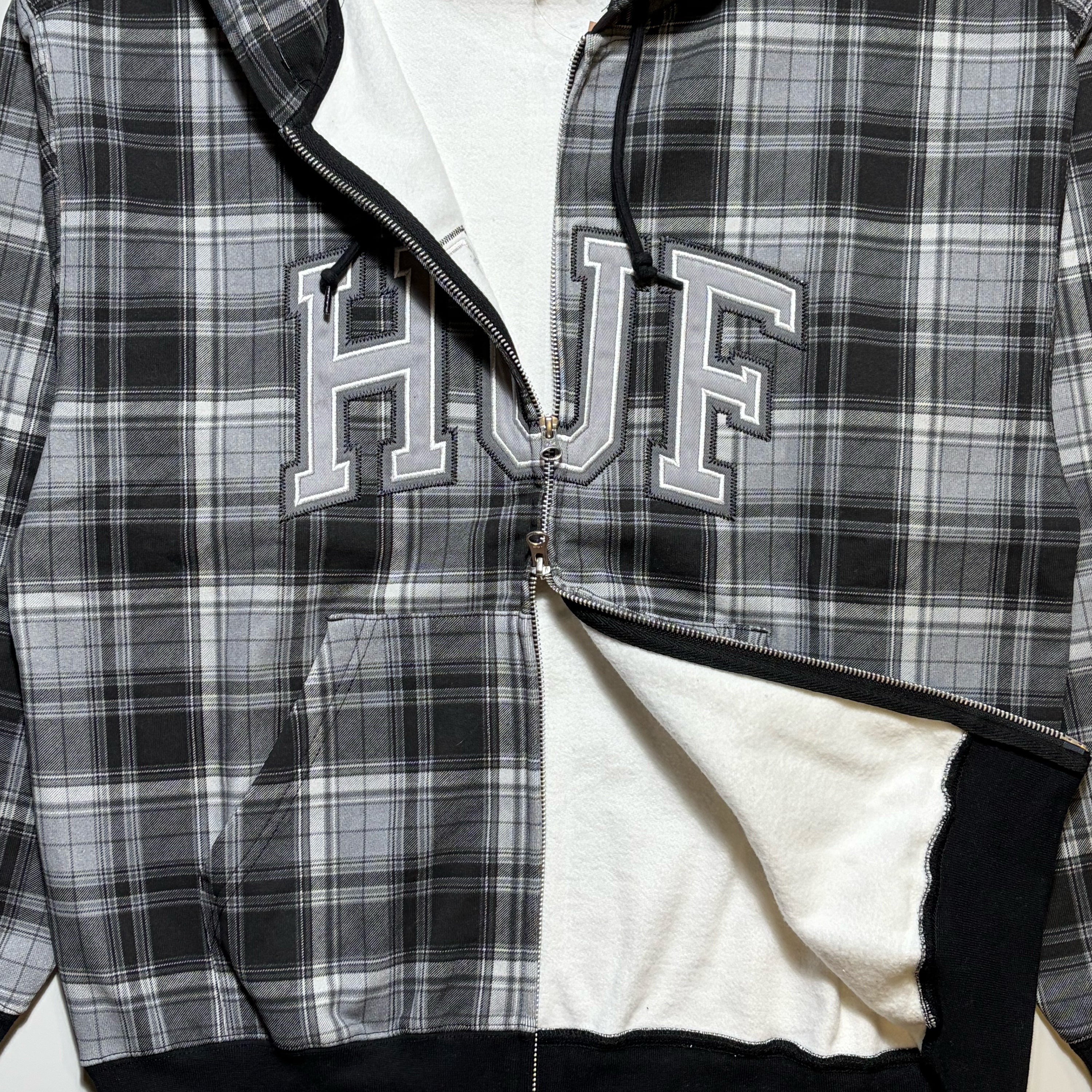 HUF double zip-up hoodie featuring a black, white, and gray plaid pattern, with 'HUF' embroidered in varsity block letters across the chest. Fleece lined.