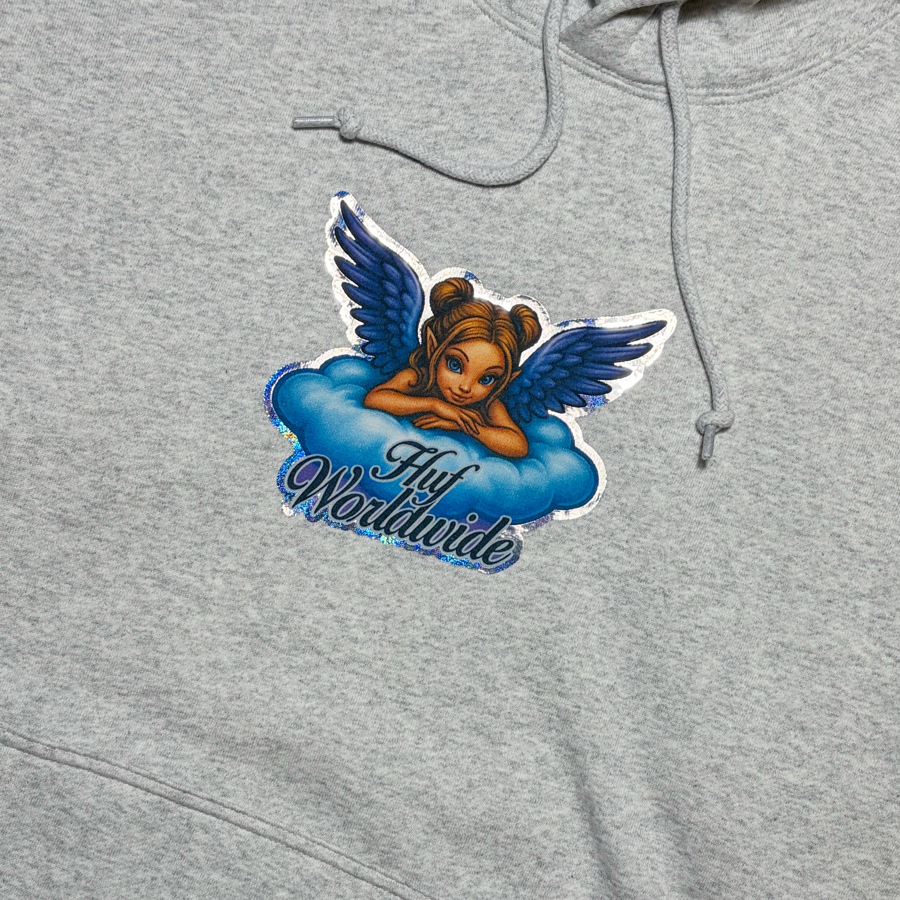 A close-up of a heather gray hoodie featuring a graphic sticker of a cartoon angel with blue wings resting on a blue cloud, with the text 'HUF Worldwide' written below.