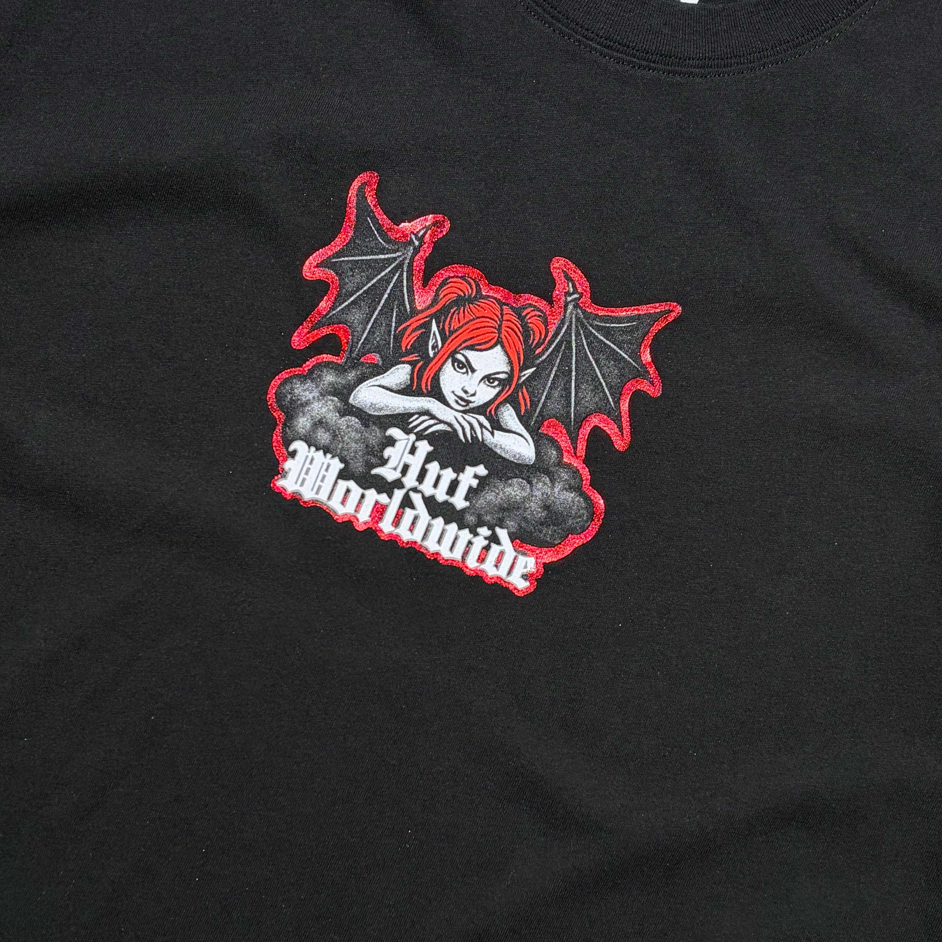 Close-up of a black t-shirt featuring a graphic print of a red-haired angel with bat wings and the text 'HUF Worldwide' in white gothic lettering.