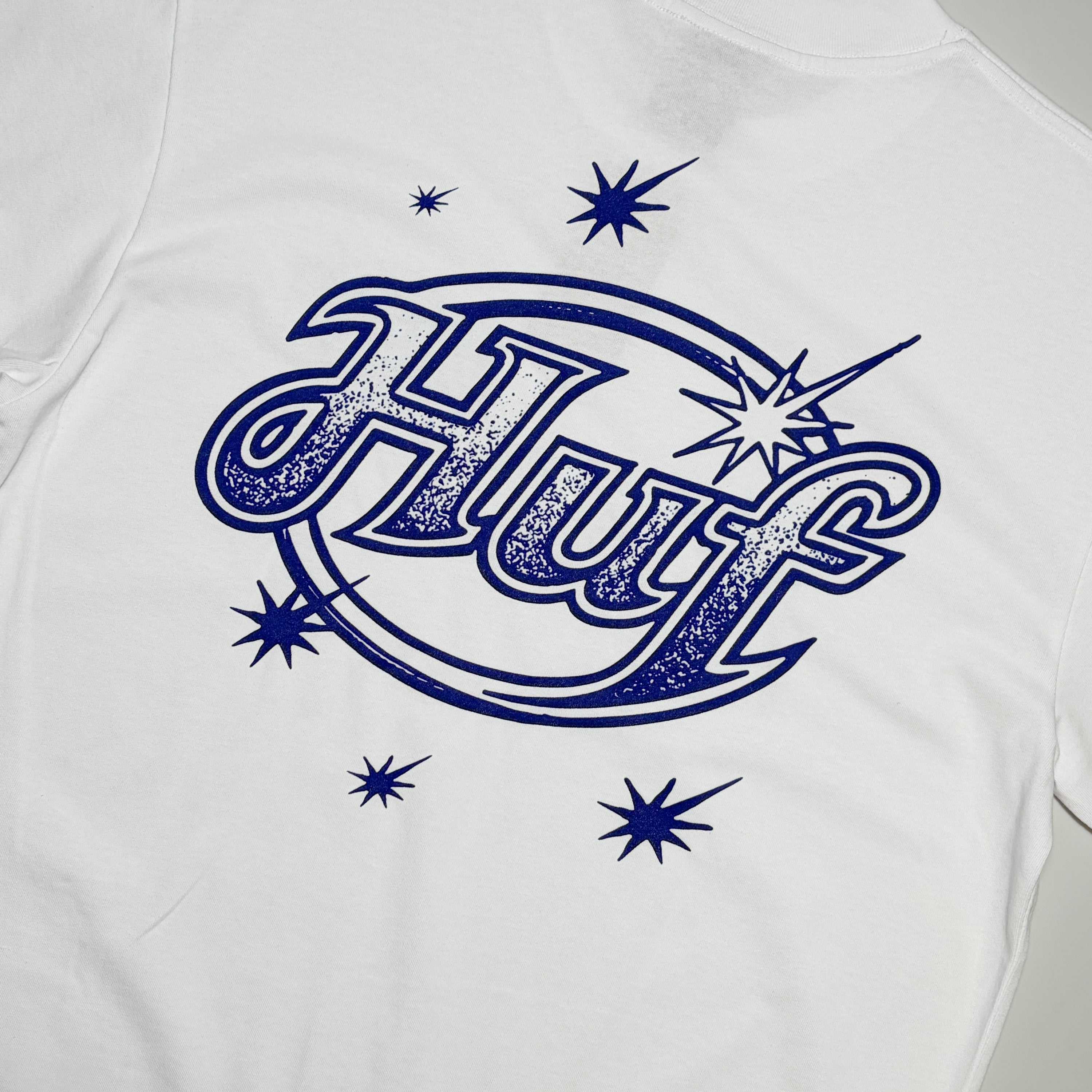 White short-sleeve t-shirt featuring a distressed navy blue graphic on the chest with the stylized text 'Huf' inside an oval, surrounded by starburst shapes.
