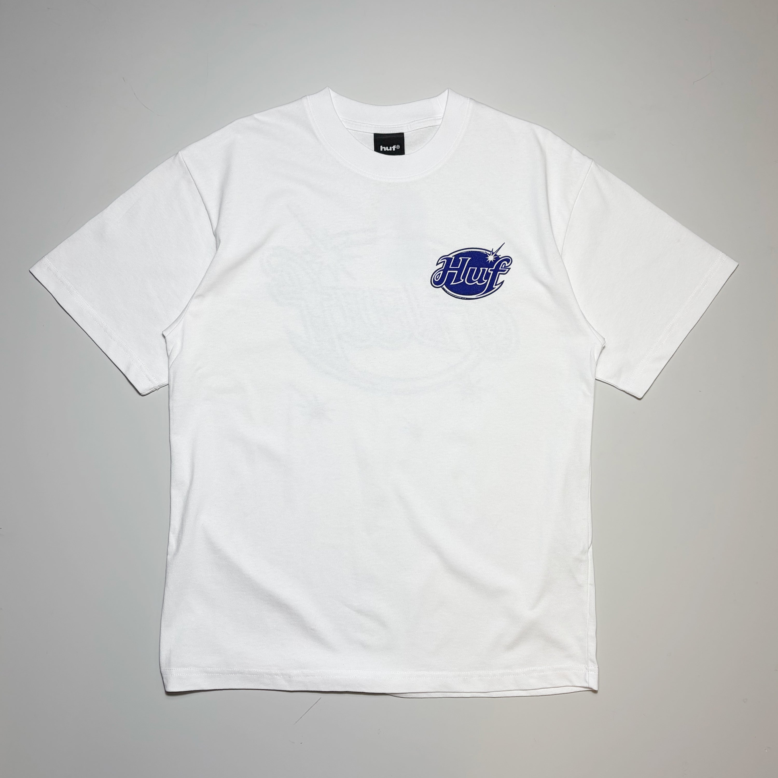 A white short-sleeve crewneck t-shirt with a blue and white 'HUF' logo graphic on the left chest, laid flat against a neutral gray background.
