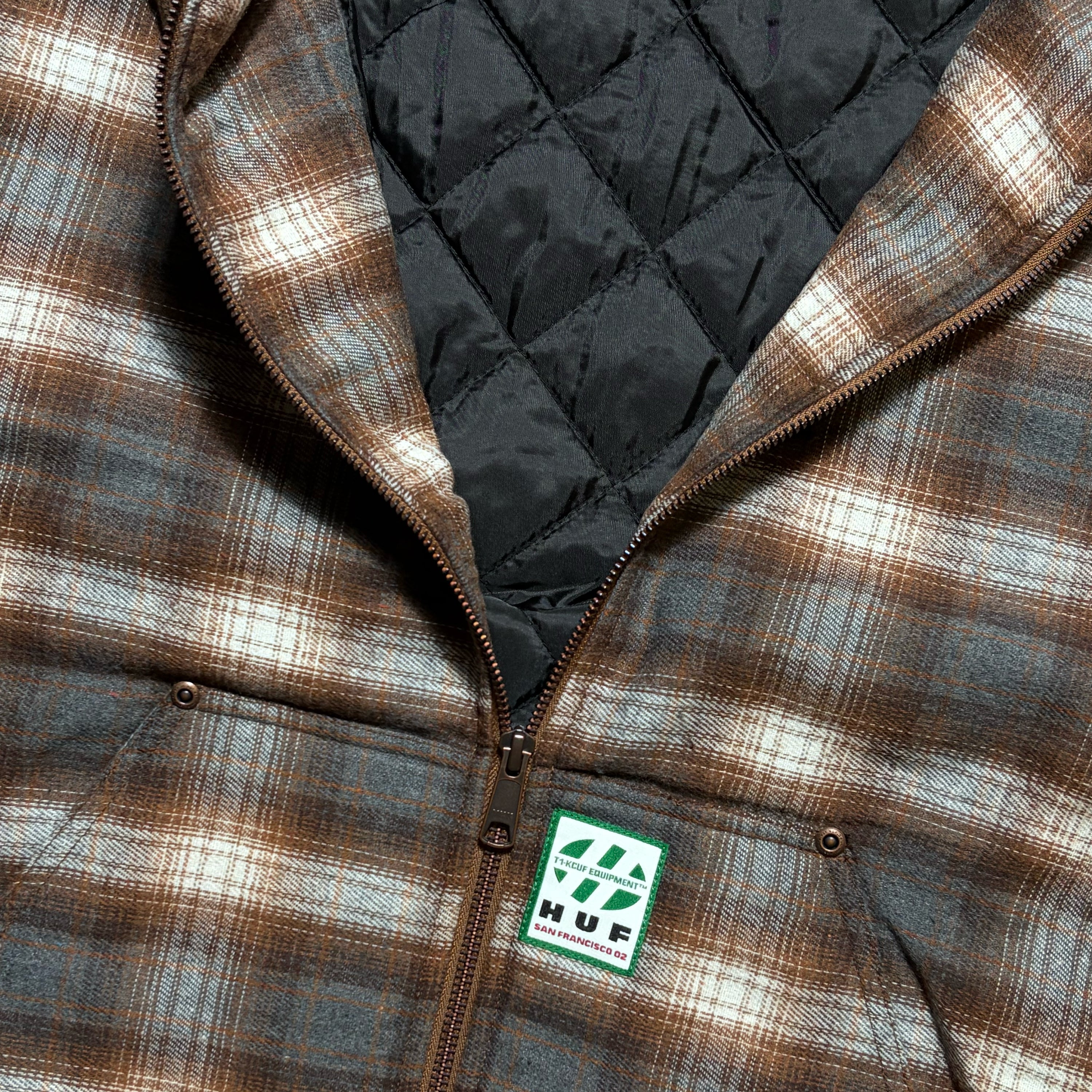 Close-up of a brown and beige plaid zip-up jacket with a black quilted lining and a visible HUF logo patch near the bottom front.