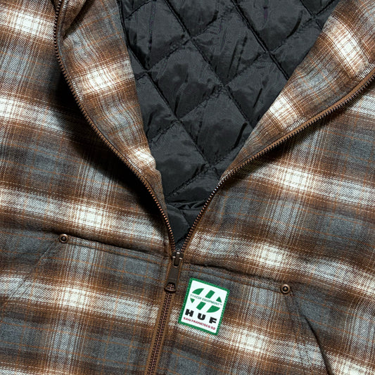 Close-up of a brown and beige plaid zip-up jacket with a black quilted lining and a visible HUF logo patch near the bottom front.