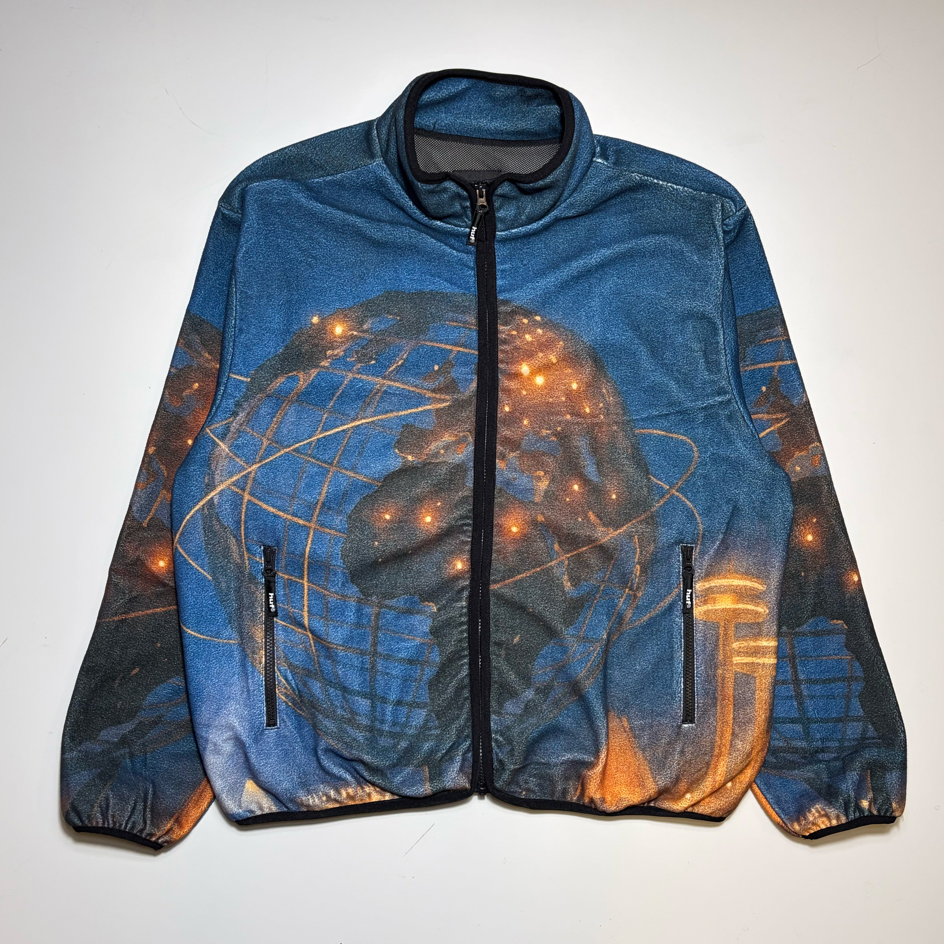 HUF Polar fleece in blue zip-up jacket with a digital printed graphic of a globe and glowing city lights, laid flat against a light background.