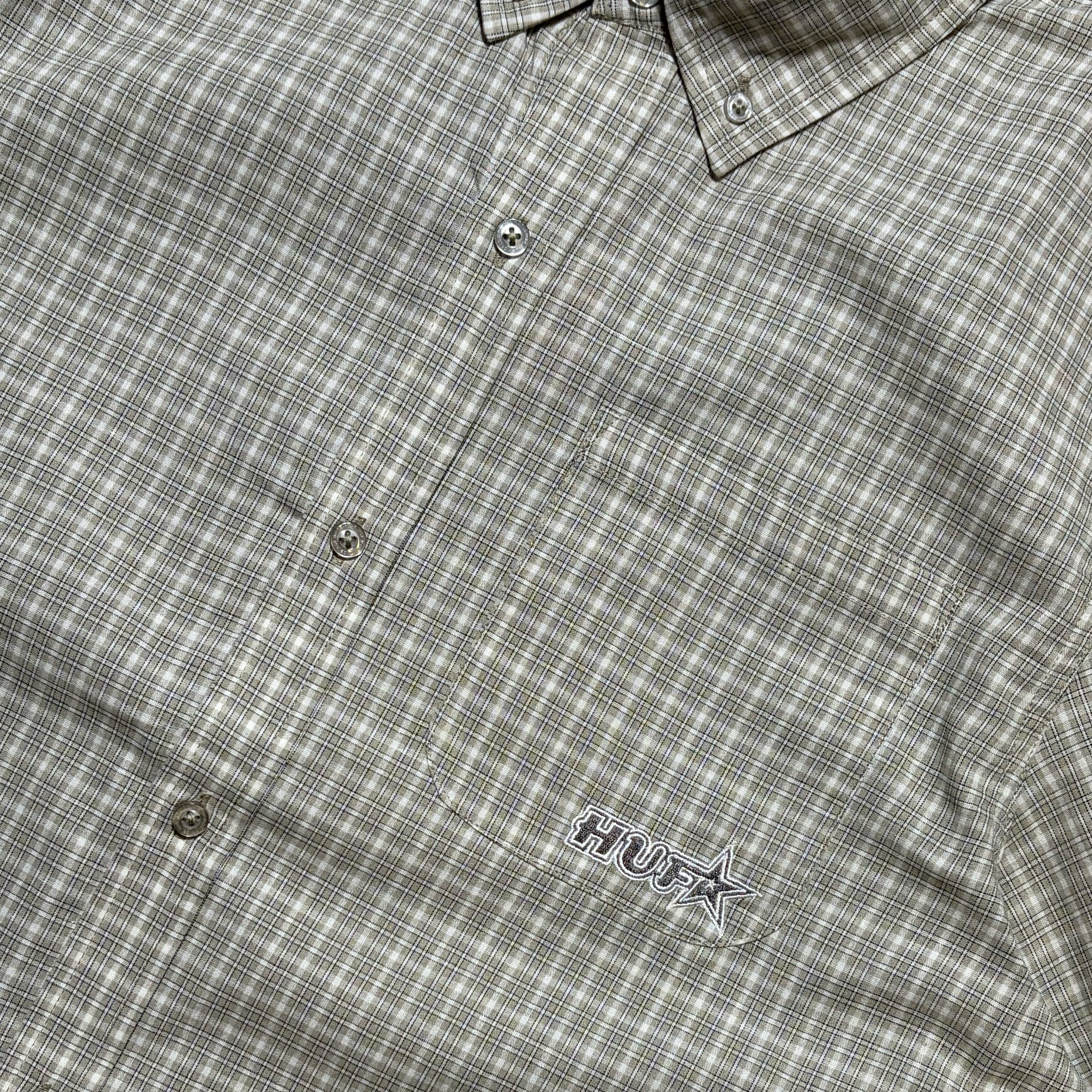 Close-up of a plaid button-down shirt in a muted gray and white pattern, featuring a small embroidered logo on the chest pocket.