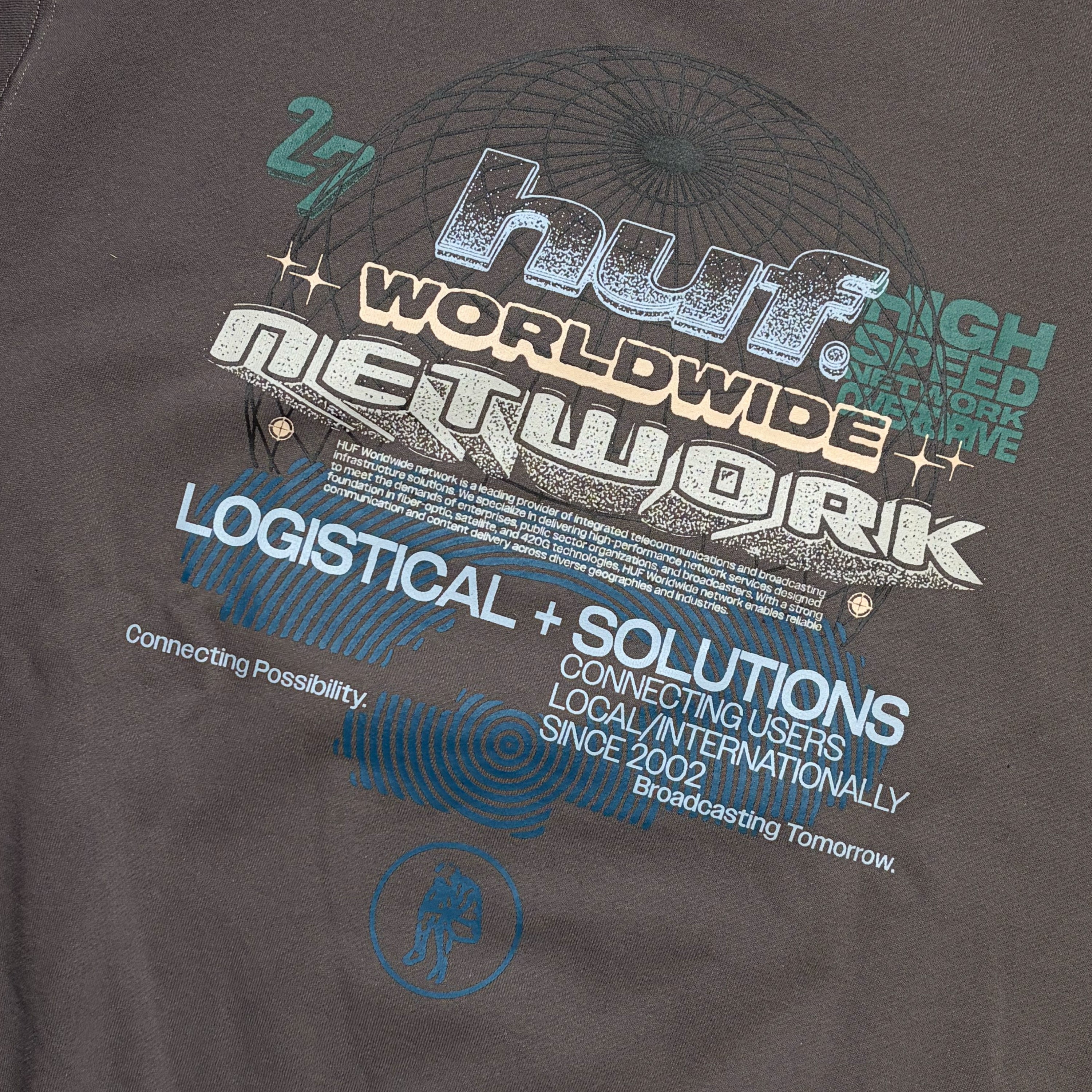Close-up of a dark gray t-shirt featuring a large graphic print for 'HUF Worldwide Network' in white, teal, and beige, with a circular globe design in the background.