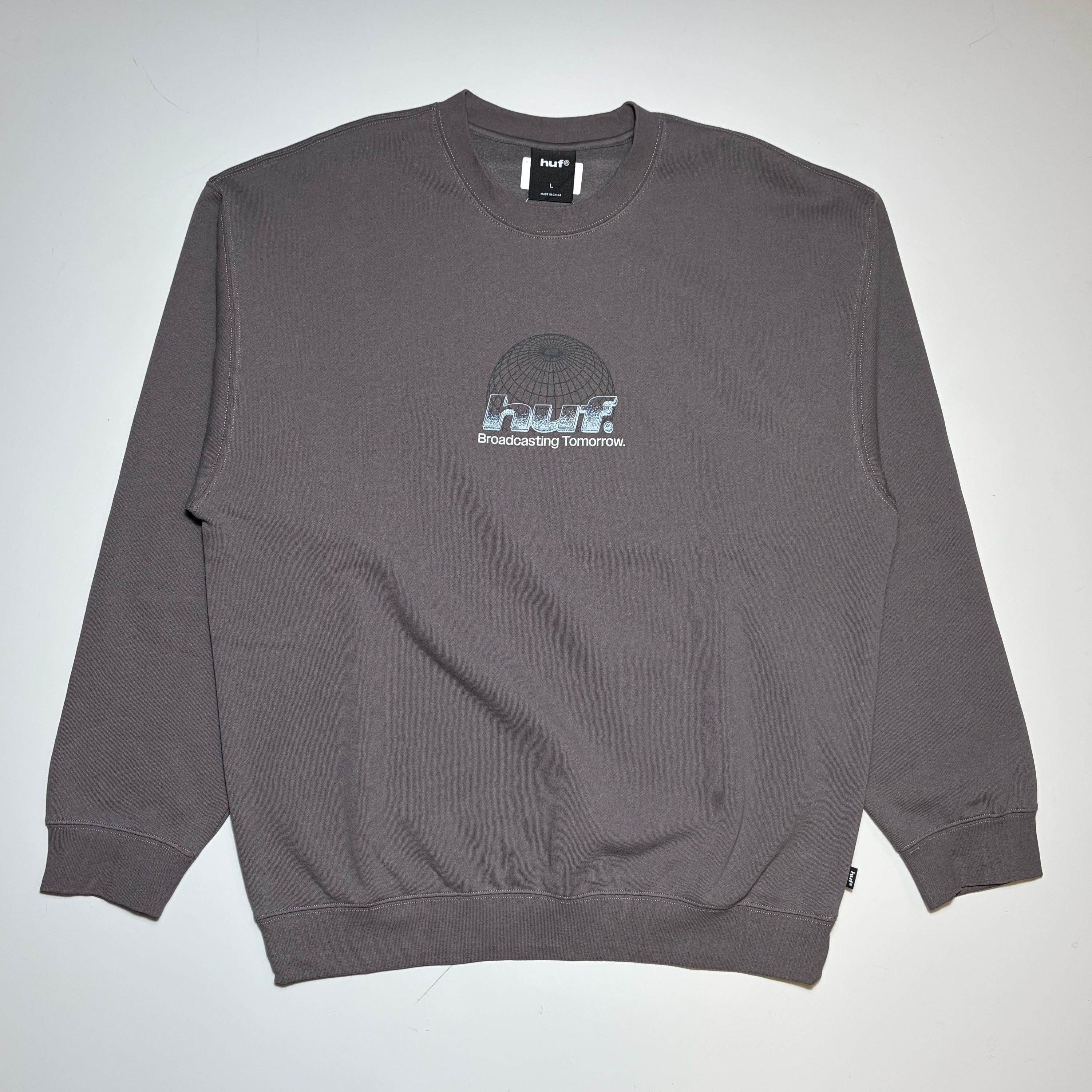 A dark gray crewneck sweatshirt with a small 'HUF' graphic on the center chest, laid flat against a light gray background.