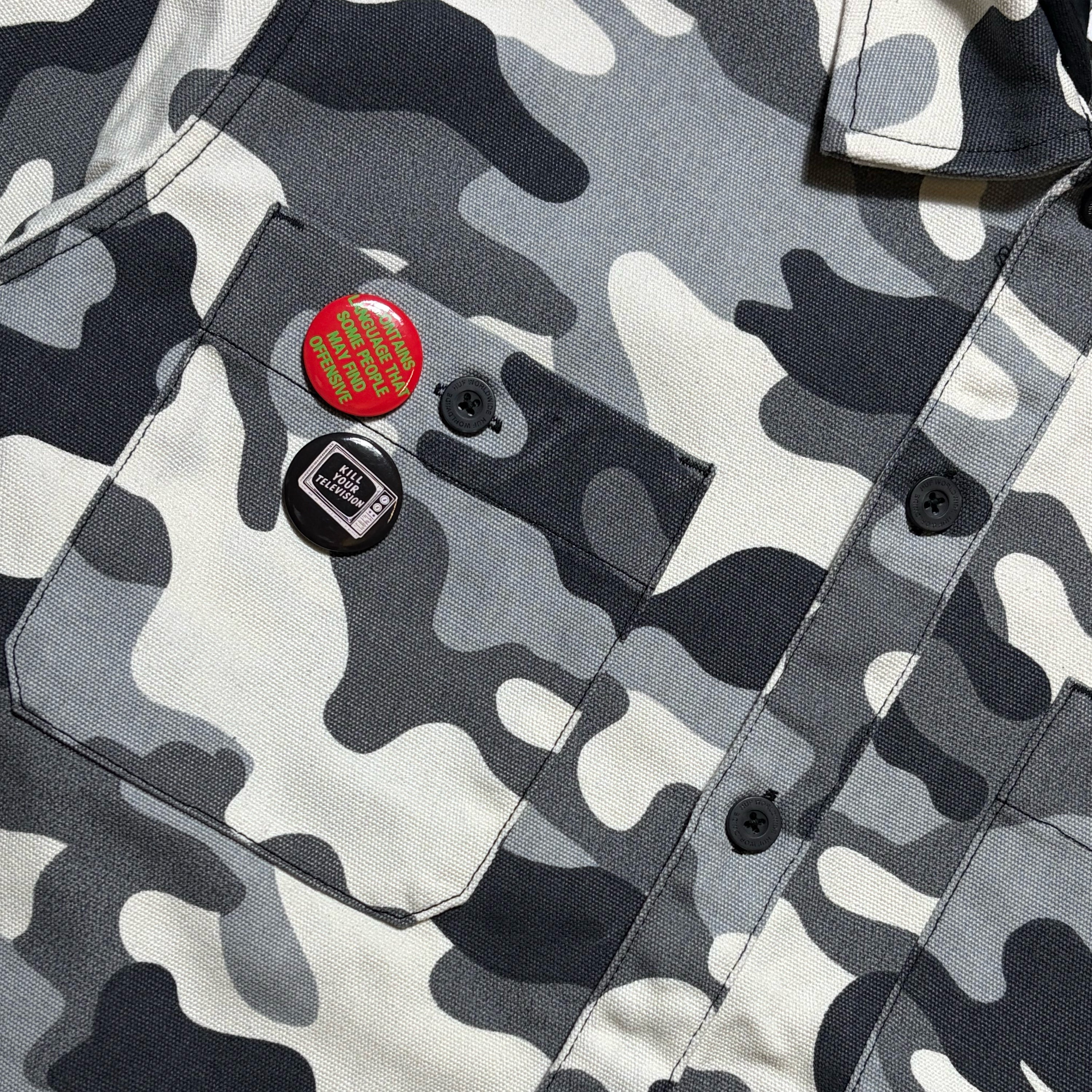 Close-up of a camouflage button-up shirt with a chest pocket, featuring two pins: one red with a gold grid pattern and one black with a white cassette graphic, on a gray, white, and black camo fabric.