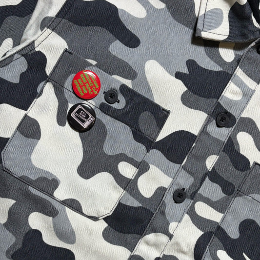 Close-up of a camouflage button-up shirt with a chest pocket, featuring two pins: one red with a gold grid pattern and one black with a white cassette graphic, on a gray, white, and black camo fabric.