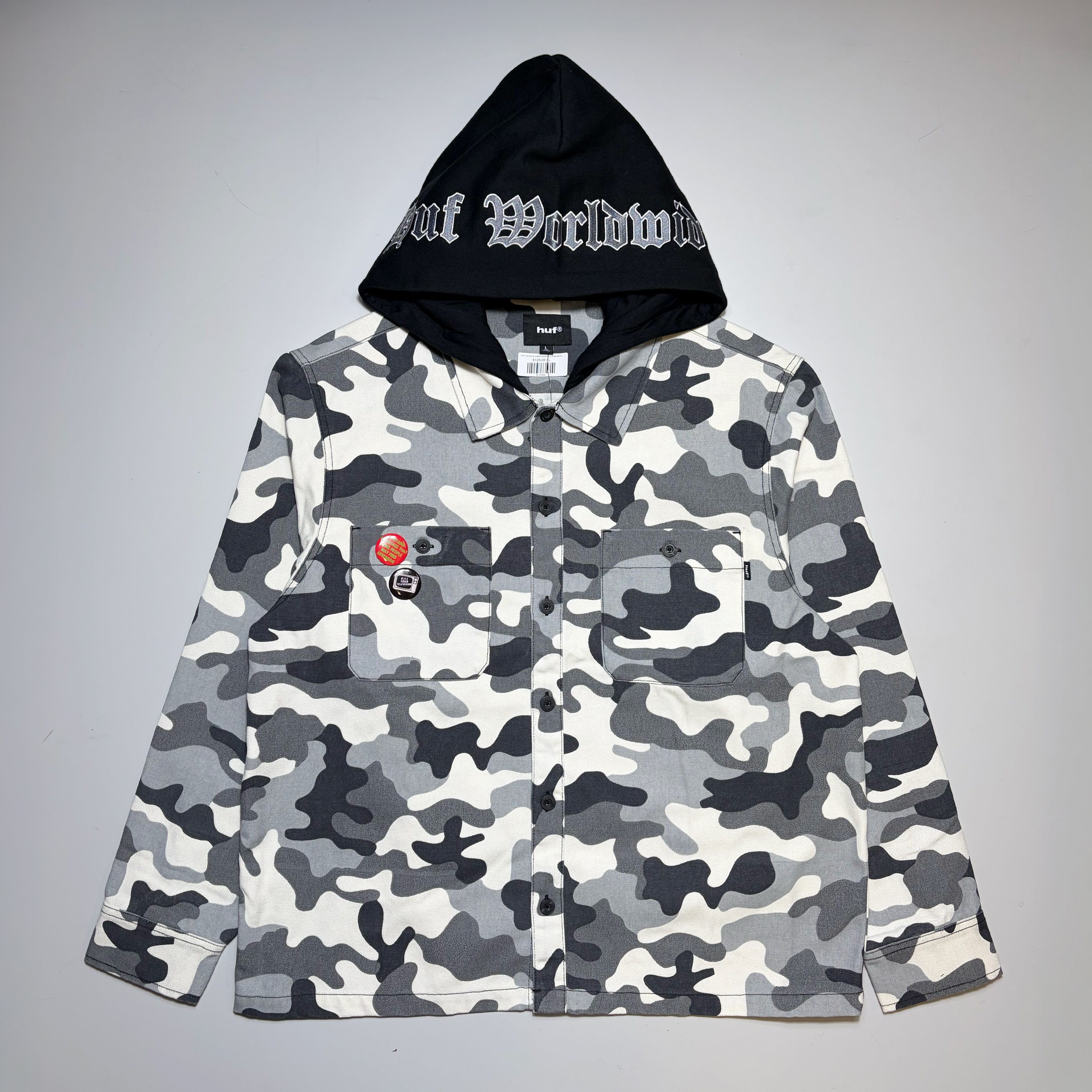 A camouflage button-up shirt jacket with a black hood featuring white Gothic lettering that reads 'HUF Worldwide', displayed flat against a neutral gray background.