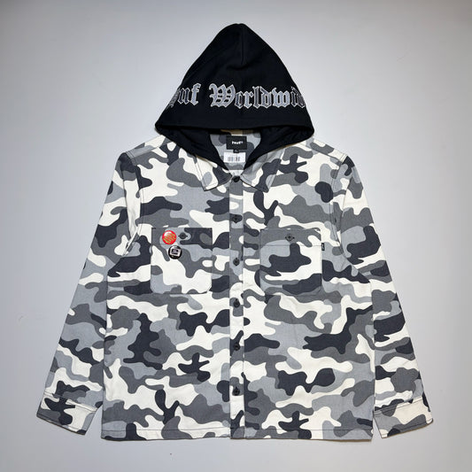 A camouflage button-up shirt jacket with a black hood featuring white Gothic lettering that reads 'HUF Worldwide', displayed flat against a neutral gray background.