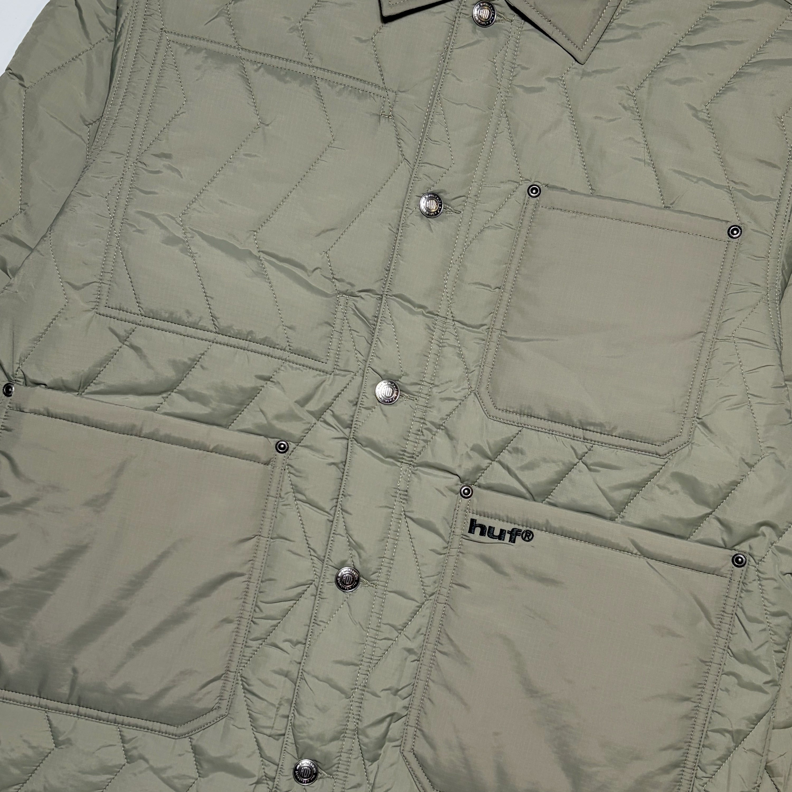 Close-up of a quilted jacket in a muted sage green color, featuring silver snap buttons, a chest pocket, and a lower patch pocket with a visible HUF logo.