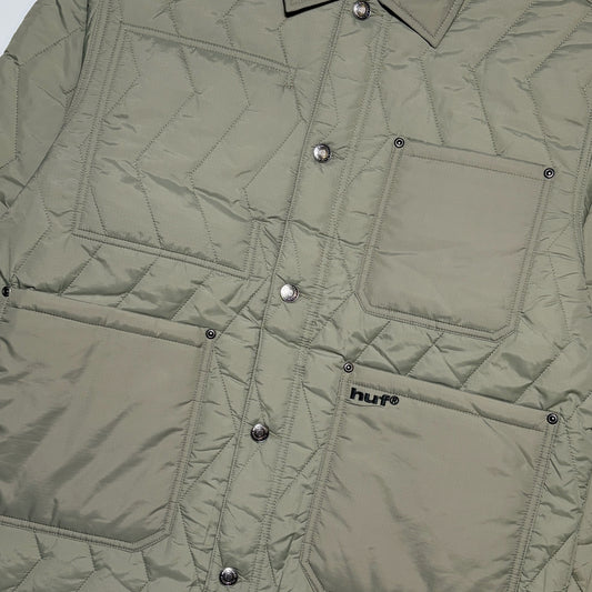 Close-up of a quilted jacket in a muted sage green color, featuring silver snap buttons, a chest pocket, and a lower patch pocket with a visible HUF logo.
