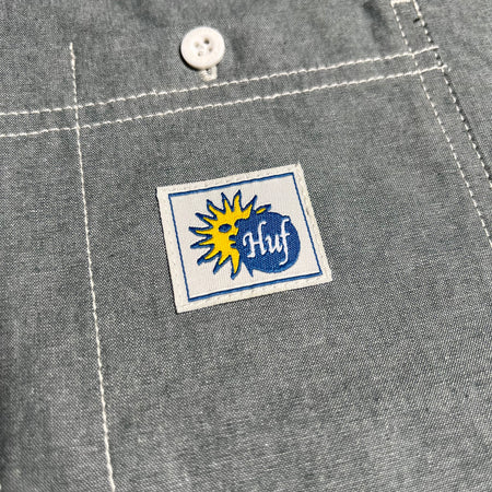 CLOSE UP IMAGE OF THE HUF ECLIPSE LOGO PATCH STITCHED ON A CHAMBRAY DRESS SHIRT CHEST POCKET