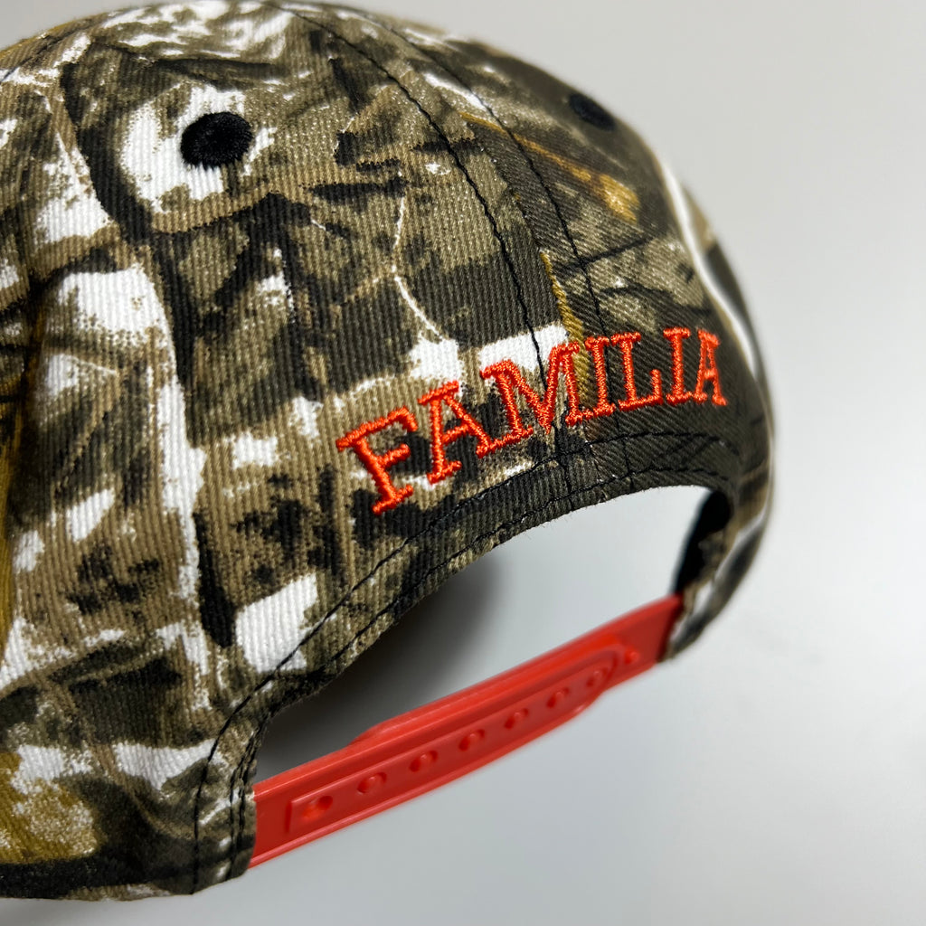 Backside of unstructured 6 panel in camo print with orange embroidered familia above orange snapback.