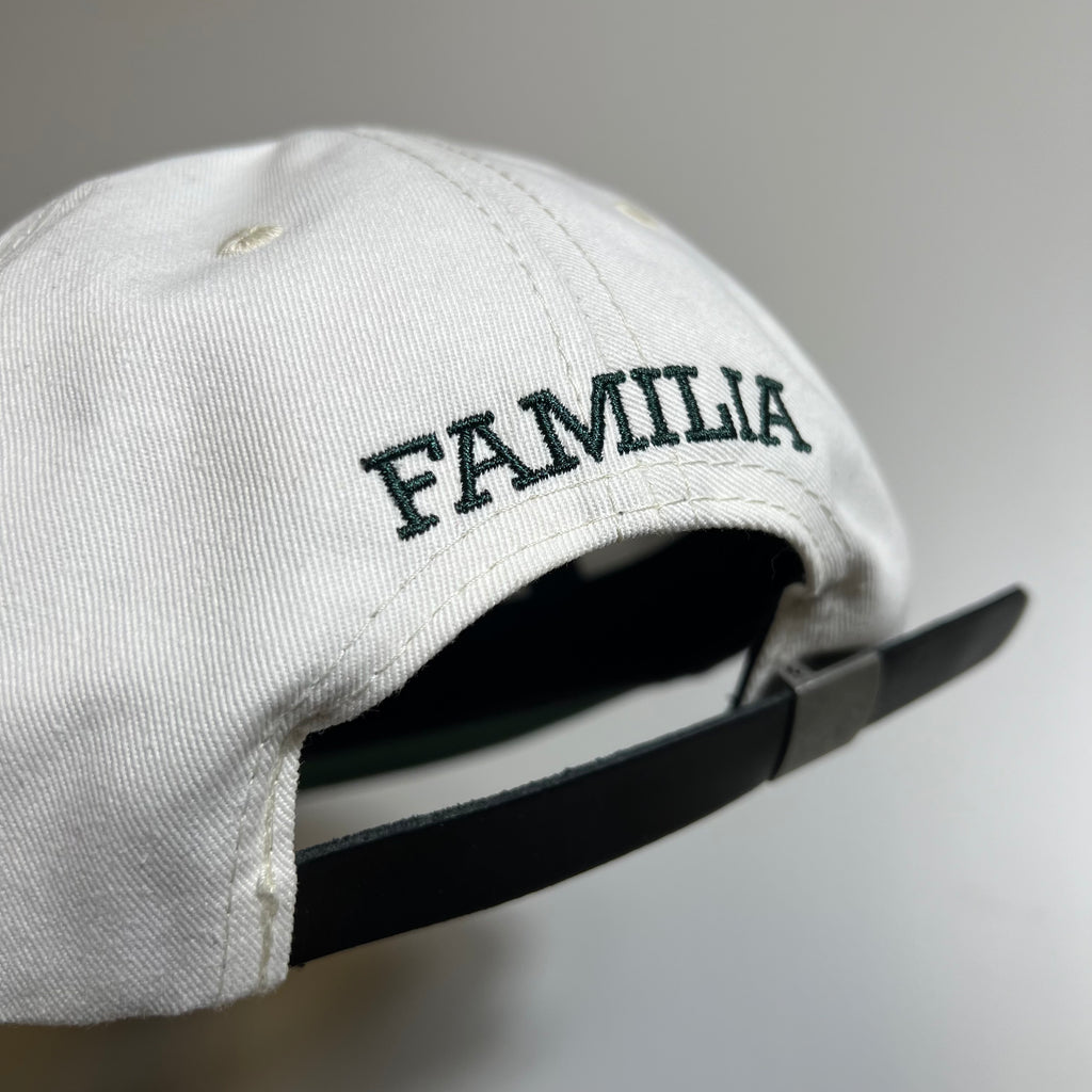 Back facing unstructured 6 panel hat in white with green familia embroidered on back above black leather strap.