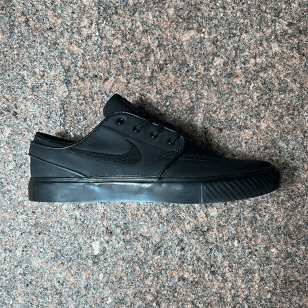 NIKE SB JANOSKI SHOE IN BLACK LEATHER, BLACK EMBROIDERED SWOOSH LOGO AND BLACK SOLE DISPLAYED ON GRANITE