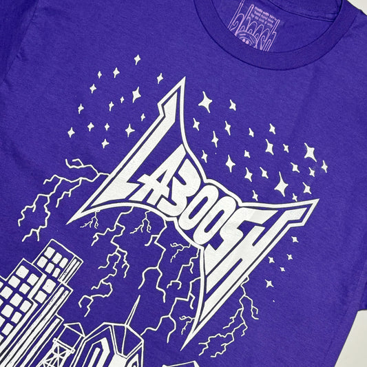 A close-up of a purple t-shirt featuring a white graphic print of the word 'LABOOSH' in a stylized metal font, surrounded by stars, lightning, and buildings.