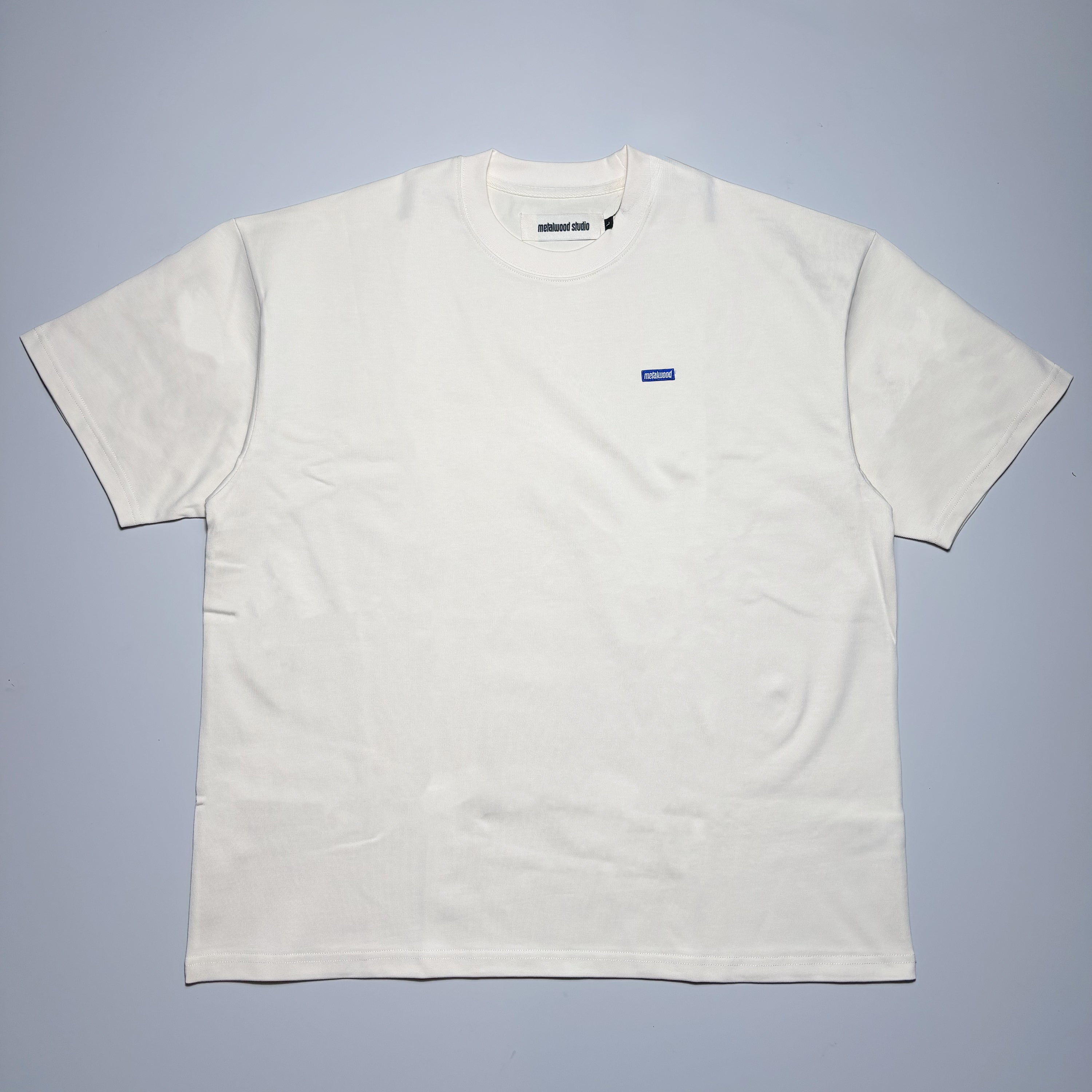 Front image of bone white tee with small blue label on chest.