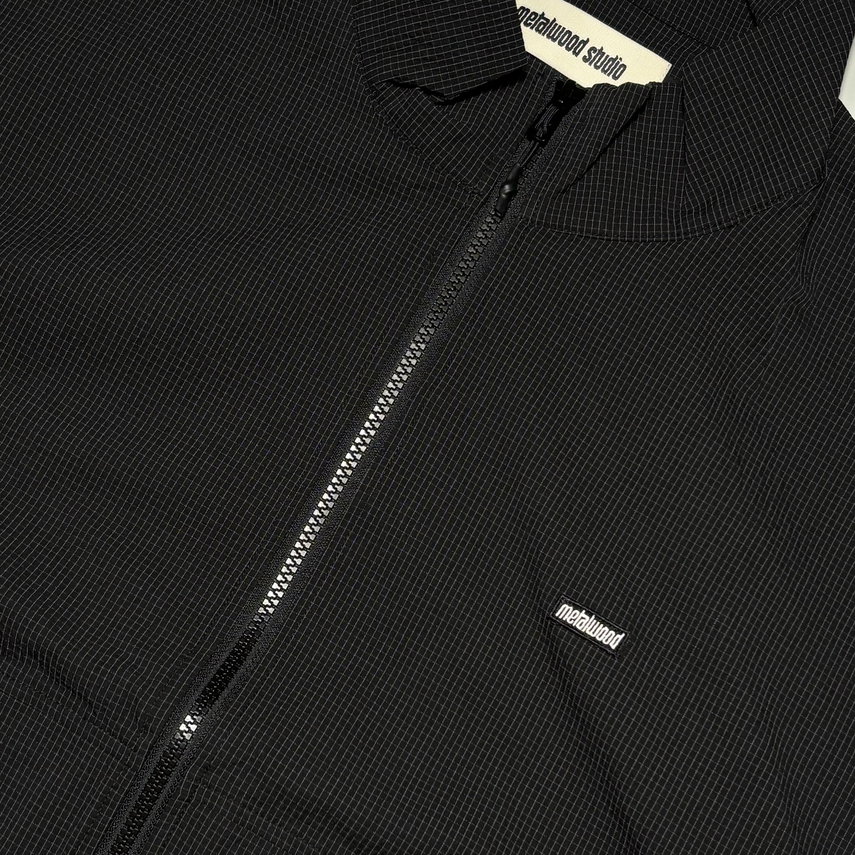 Close detail on black micro plaid track jacket with black woven label on chest.