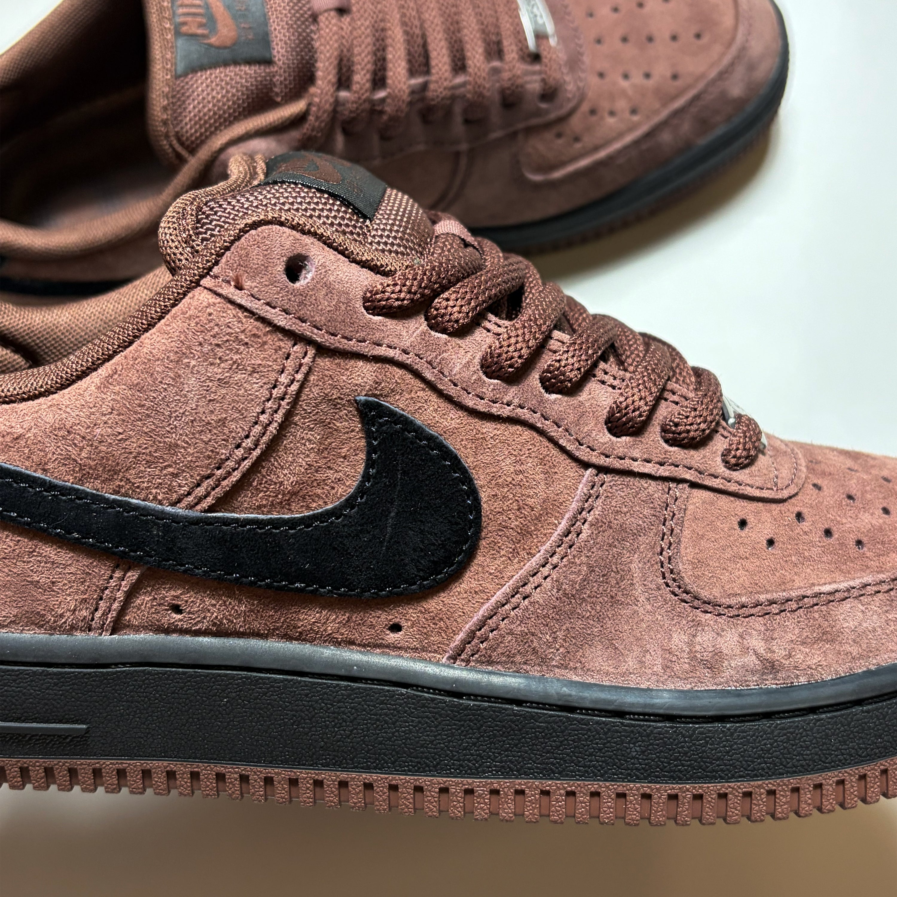 A close-up of a brown suede Nike SB Air Force 1 Low sneaker with a black Swoosh and black rubber sole, with a second matching shoe blurred in the background.
