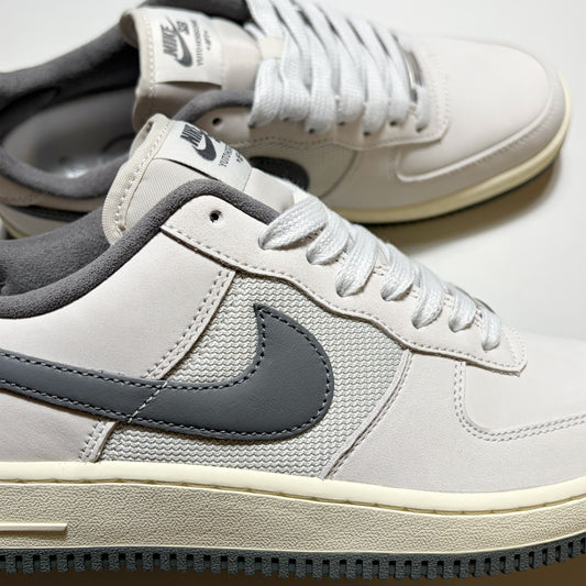 A close-up of a Nike SB Air Force 1 Low sneaker in light gray and dark gray, with a cream midsole and white laces, shown alongside another shoe in the background.