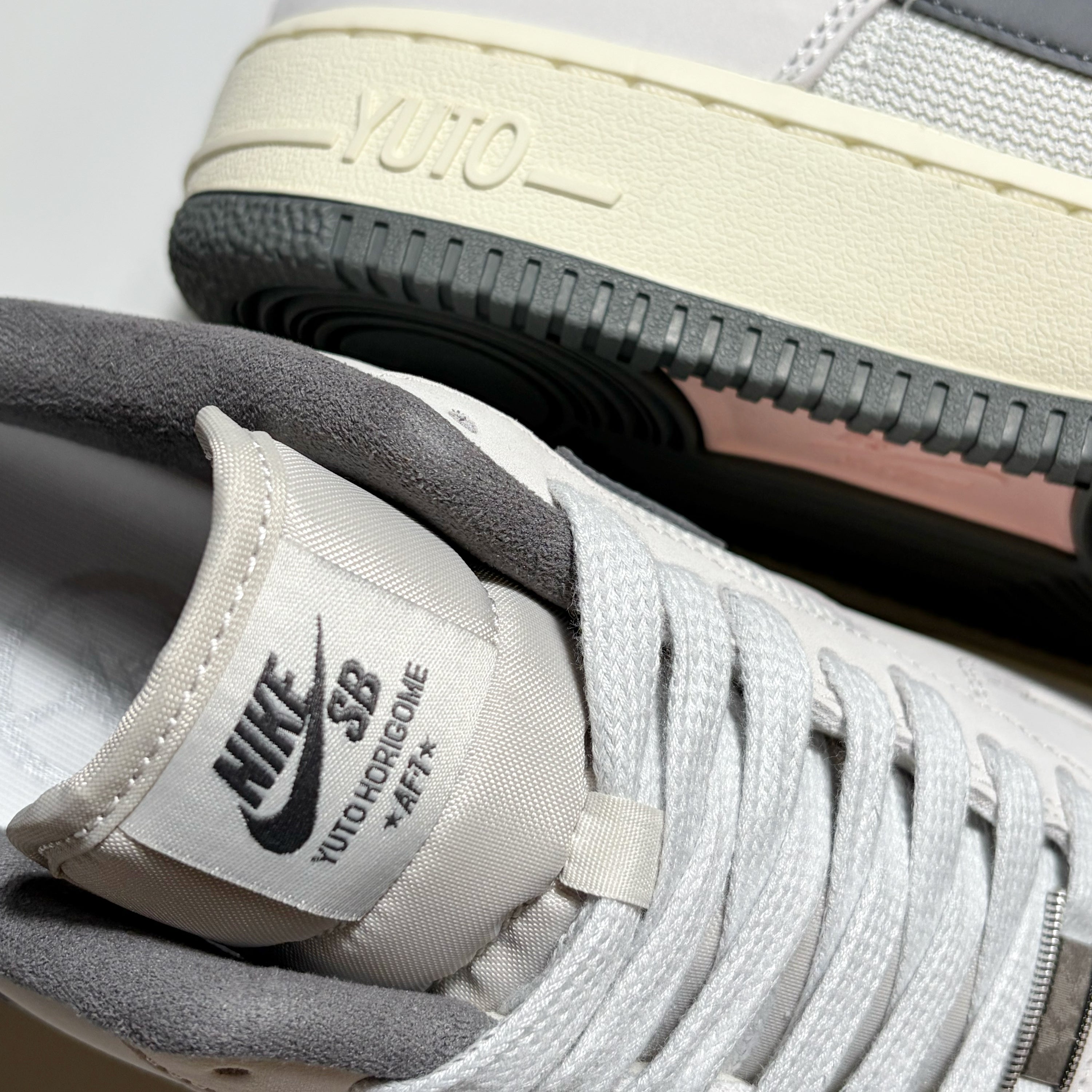 Close-up of a Nike SB Yuto Hongoime x Air Force 1 Low sneaker in light gray and off-white, showing the tongue label, laces, and the outsole with 'YUTO' branding.
