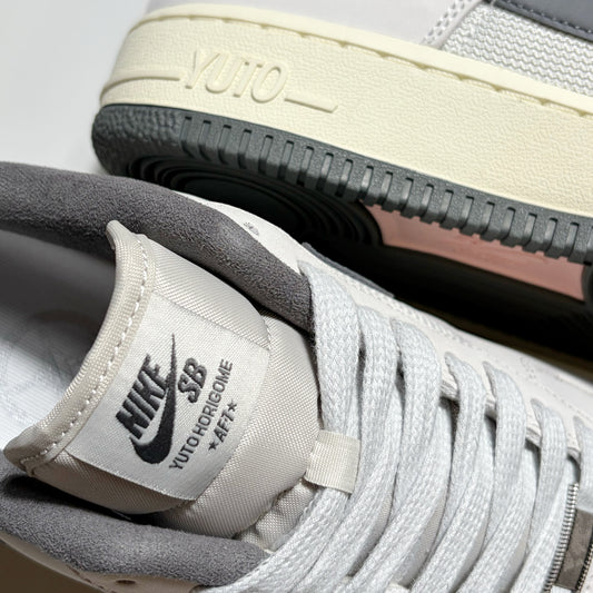 Close-up of a Nike SB Yuto Hongoime x Air Force 1 Low sneaker in light gray and off-white, showing the tongue label, laces, and the outsole with 'YUTO' branding.