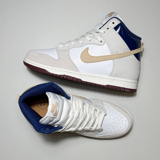 A pair of Nike SB Dunk High Pro sneakers shown from the side and top on a light gray background. The shoes feature a white leather and suede upper, a beige Swoosh, a navy blue ankle collar, and a dark burgundy outsole.