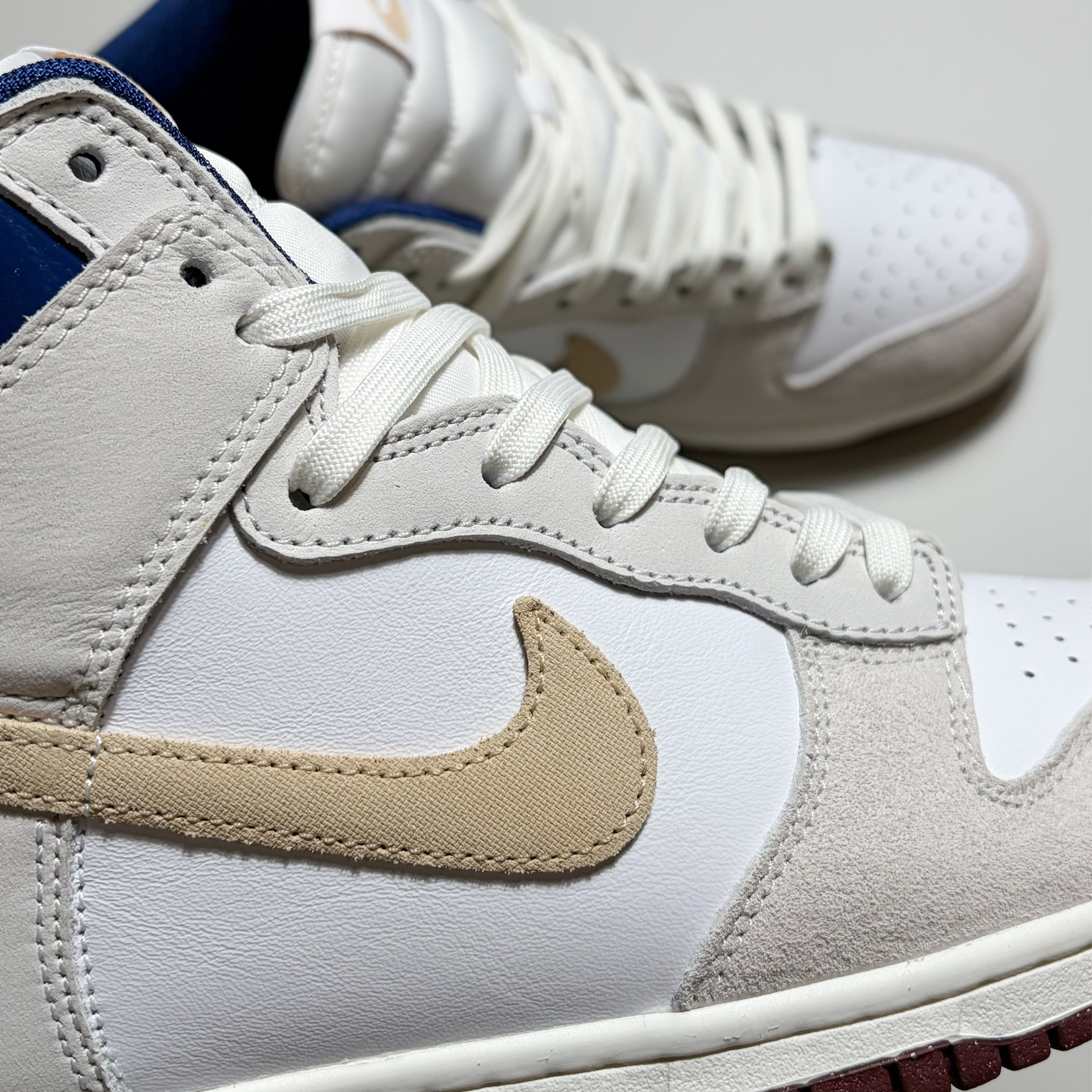 A close-up of a Nike SB high-top dunk sneaker in white leather and suede with a beige Swoosh, white laces, and a blue collar lining, with a second shoe blurred in the background.