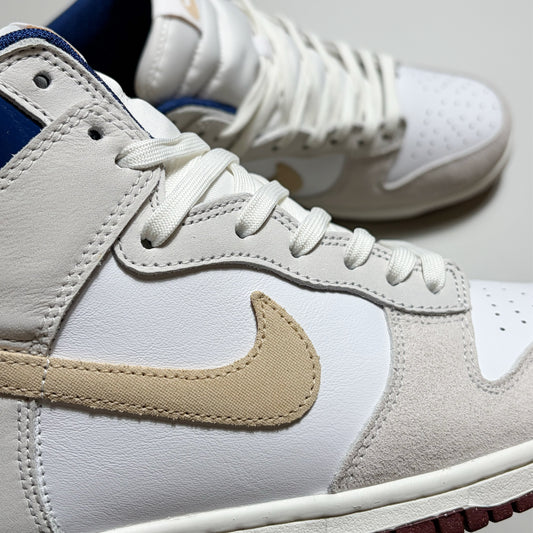 A close-up of a Nike SB high-top dunk sneaker in white leather and suede with a beige Swoosh, white laces, and a blue collar lining, with a second shoe blurred in the background.