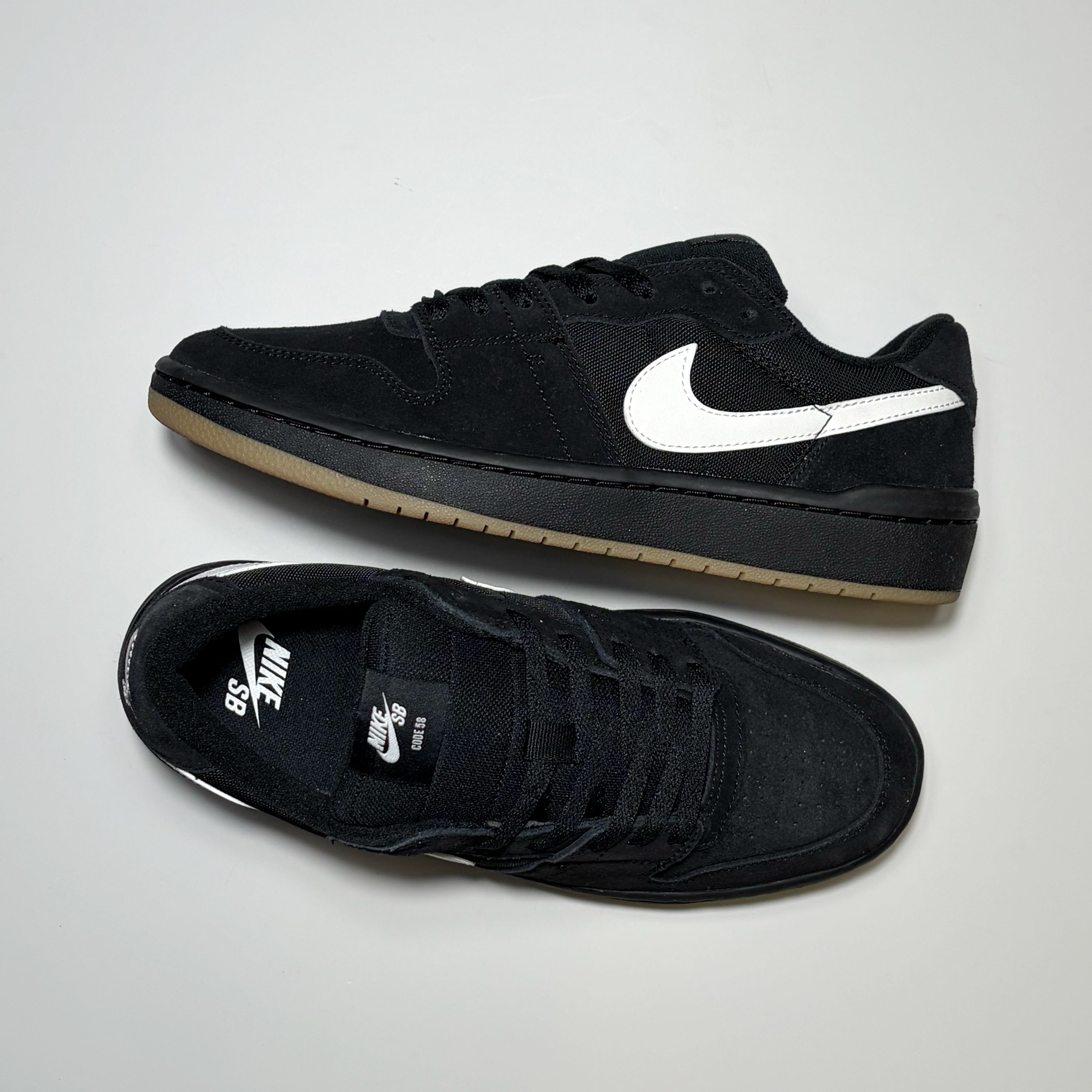 A pair of Nike SB Code 58 Low sneakers in black suede, featuring a white Swoosh on the side and a gum-colored rubber sole, displayed on a light gray background.