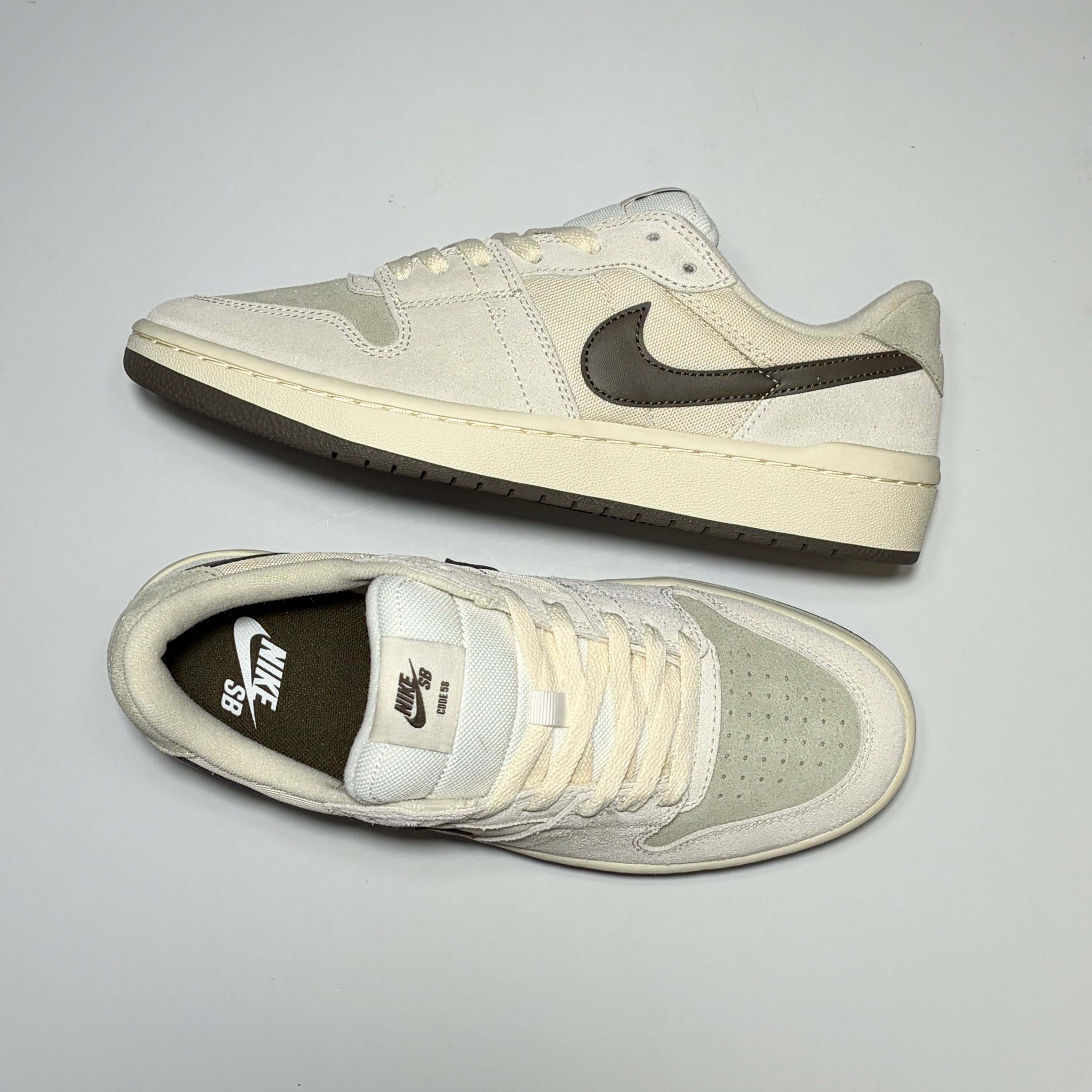 A pair of Nike SB Code 58 Low sneakers in off-white suede and leather with dark brown accents, including the Swoosh, outsole, and interior lining, displayed on a light gray background.