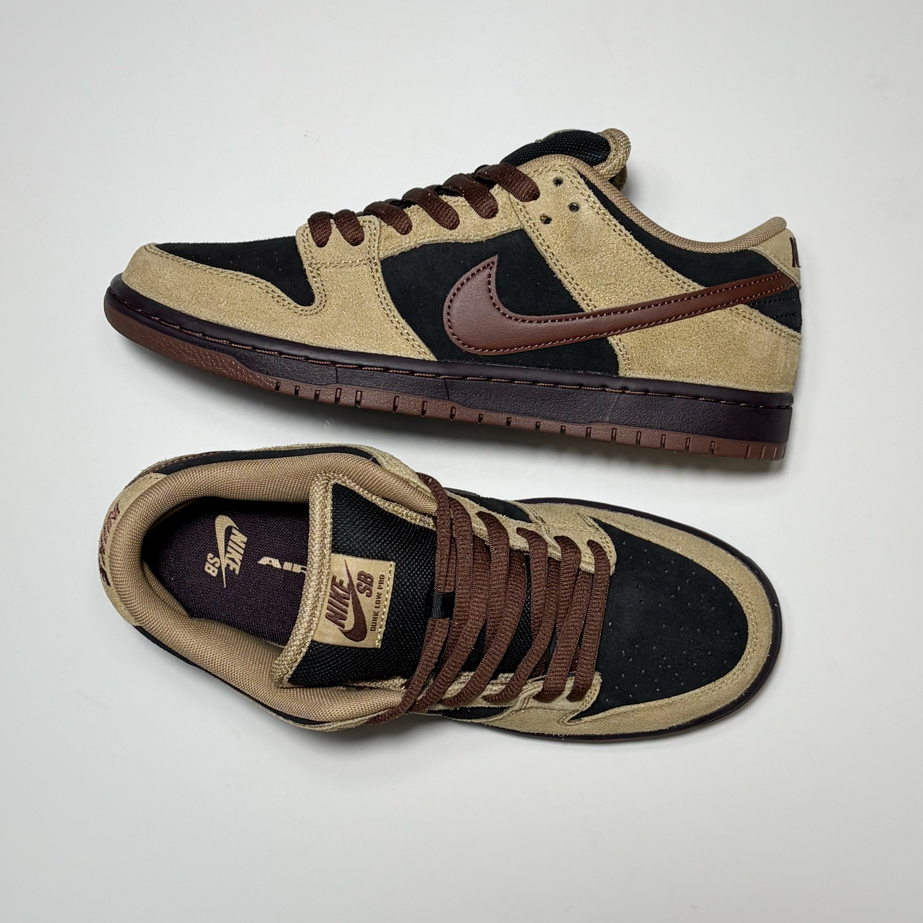 A pair of Nike SB Dunk Low sneakers in a beige, black, and brown colorway, displayed on a clean white background with one shoe showing the side profile and the other showing the top and interior.