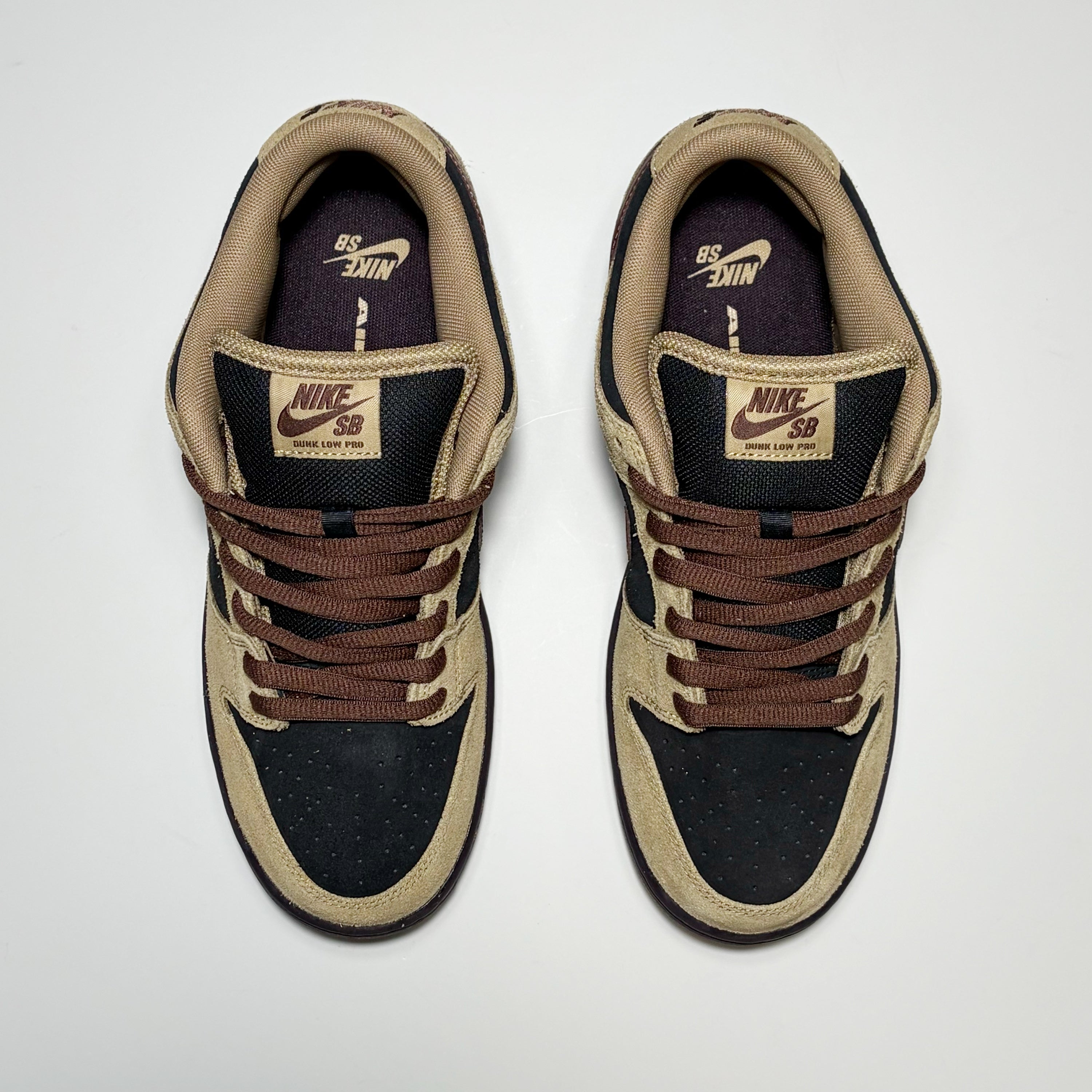 A pair of Nike SB Dunk Low Pro sneakers shown from a top-down view on a light gray background. The shoes feature a black suede upper with tan overlays, brown laces, and a dark purple Nike lining.