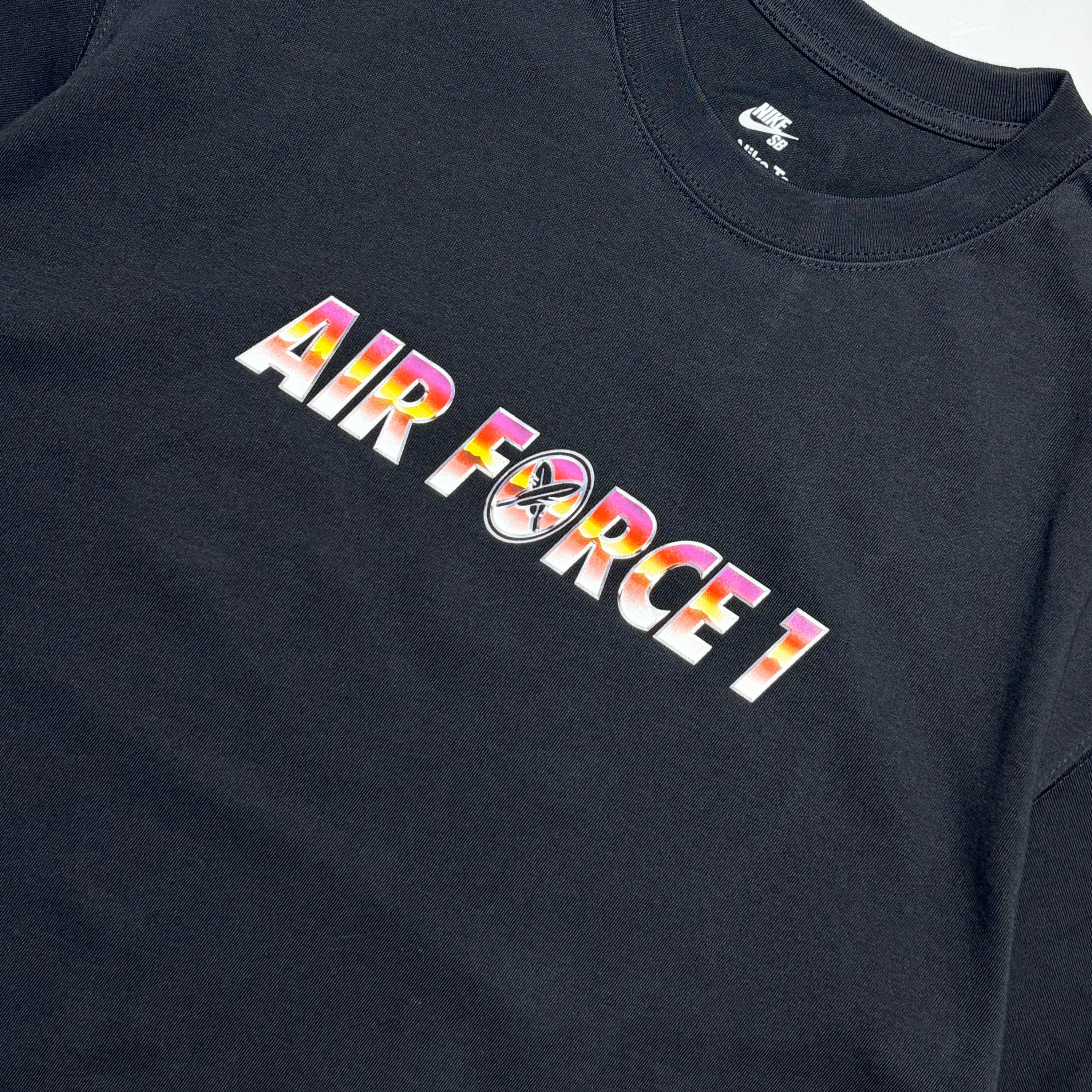 A close-up of a black Nike Air Force 1 t-shirt featuring a colorful graphic print on the chest that reads 'AIR FORCE 1' with a circular logo in the center, set against a plain white background.