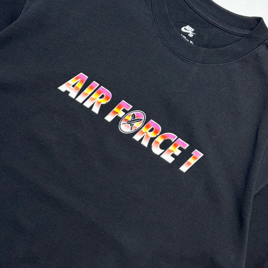 A close-up of a black Nike Air Force 1 t-shirt featuring a colorful graphic print on the chest that reads 'AIR FORCE 1' with a circular logo in the center, set against a plain white background.