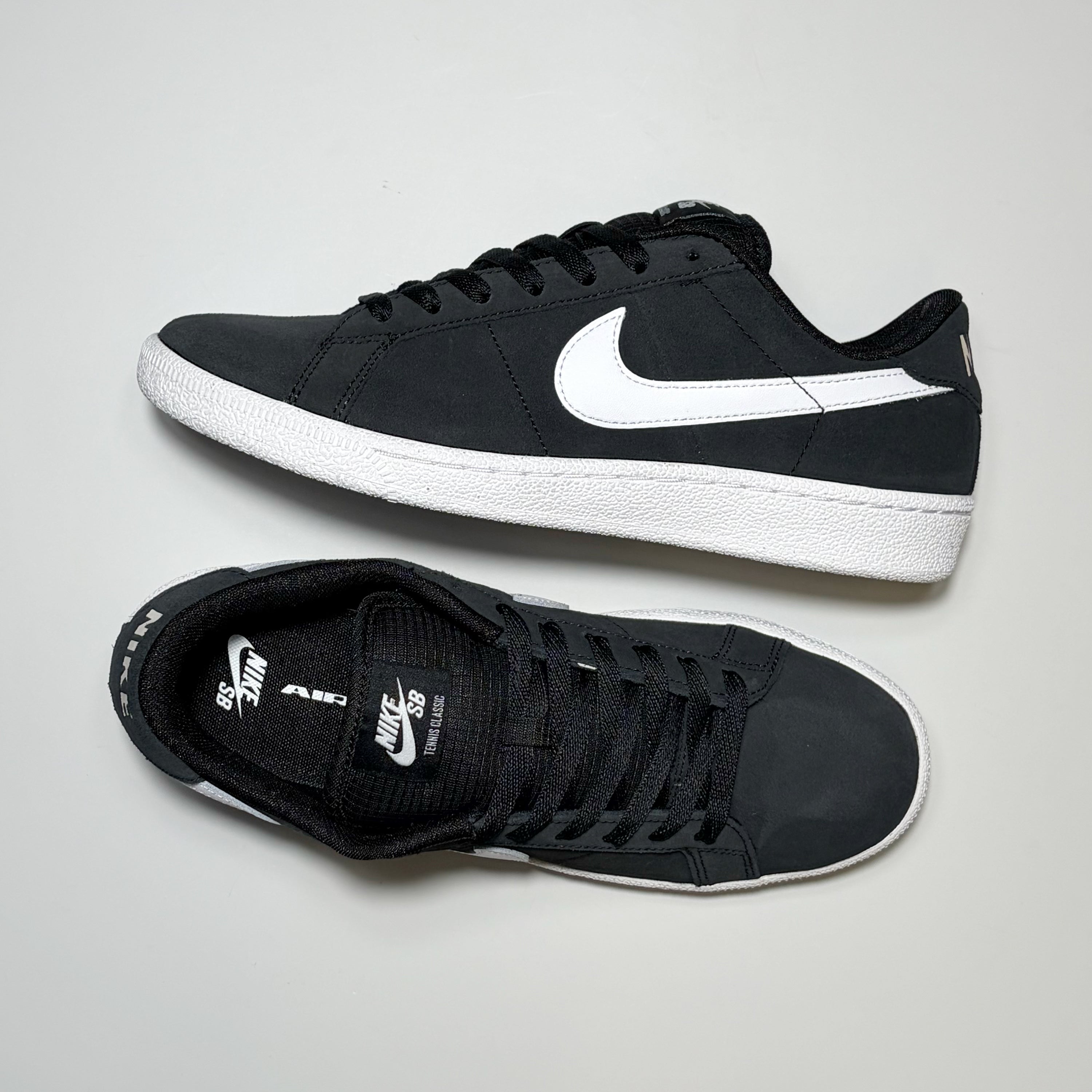 A pair of Nike SB Zoom Tennis Classic Low sneakers in black and white, displayed from side and top-down angles on a light gray background.