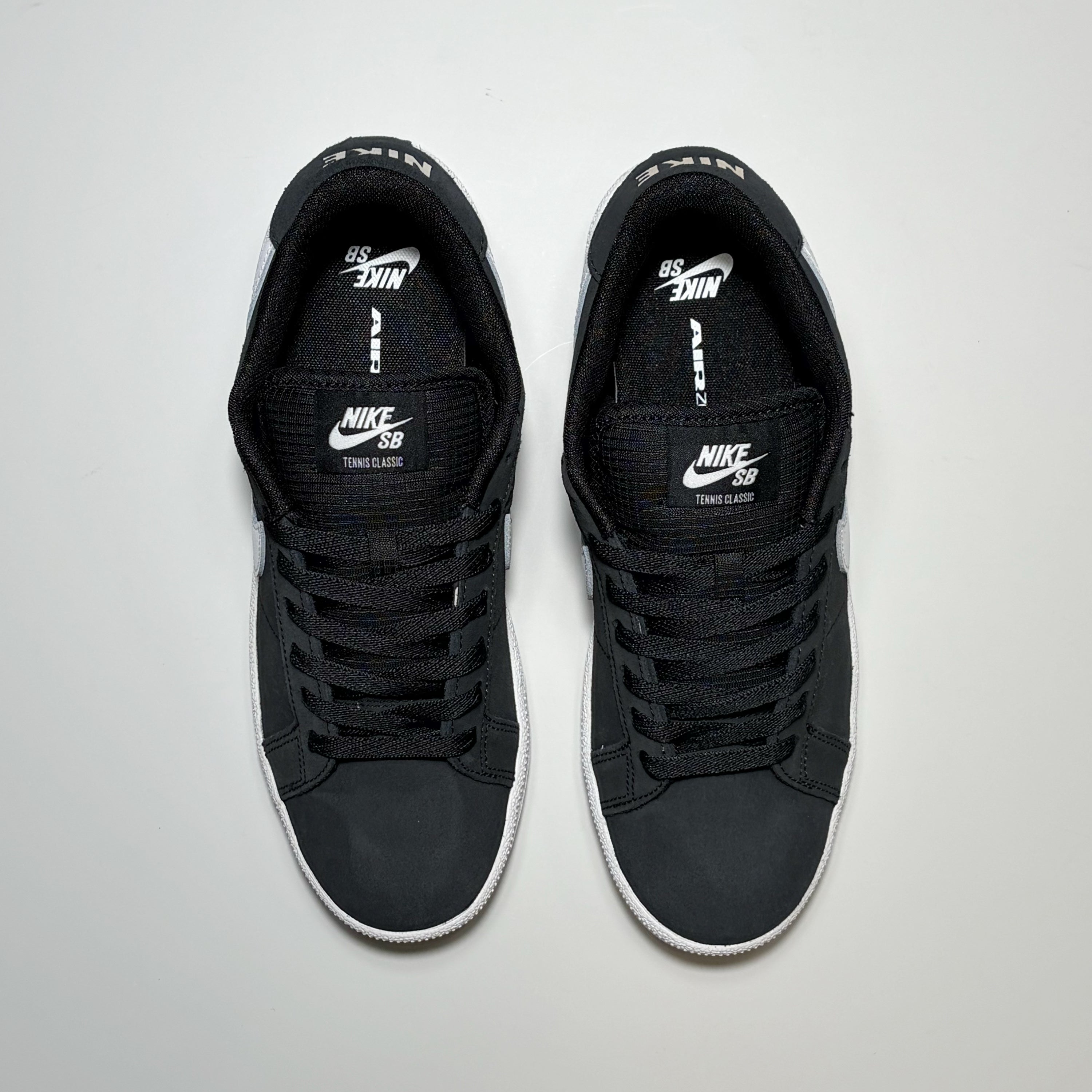 A pair of Nike SB Tennis Classic Low sneakers in black with white accents, shown from a top-down view on a light gray background.